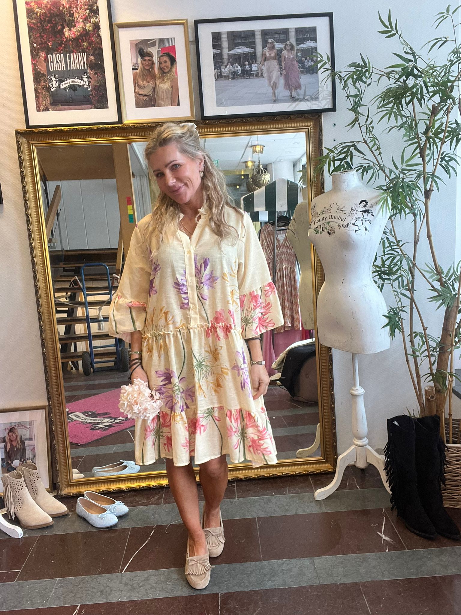 Leende kvinna står framför en stor guldramsspegel i en butik, iklädd ljus klänning med blommönster och loafers. Runt henne syns skor, en provdocka med texten “Fanny Michel” samt inramade foton, bland annat “CASA FANNY”.