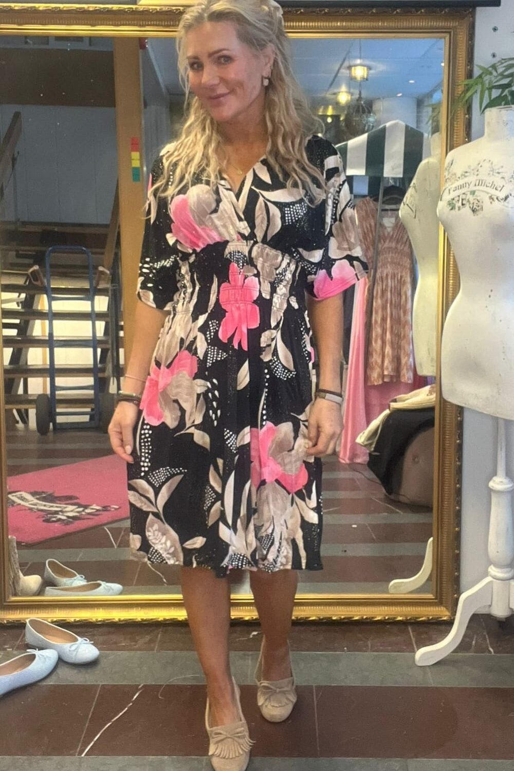 Kvinna står framför en stor guldramsspegel och poserar i en svart klänning med stora rosa och beige blommor samt ljusa loafers. Miljön liknar en butik eller ateljé, med klädställning och provdocka bredvid.