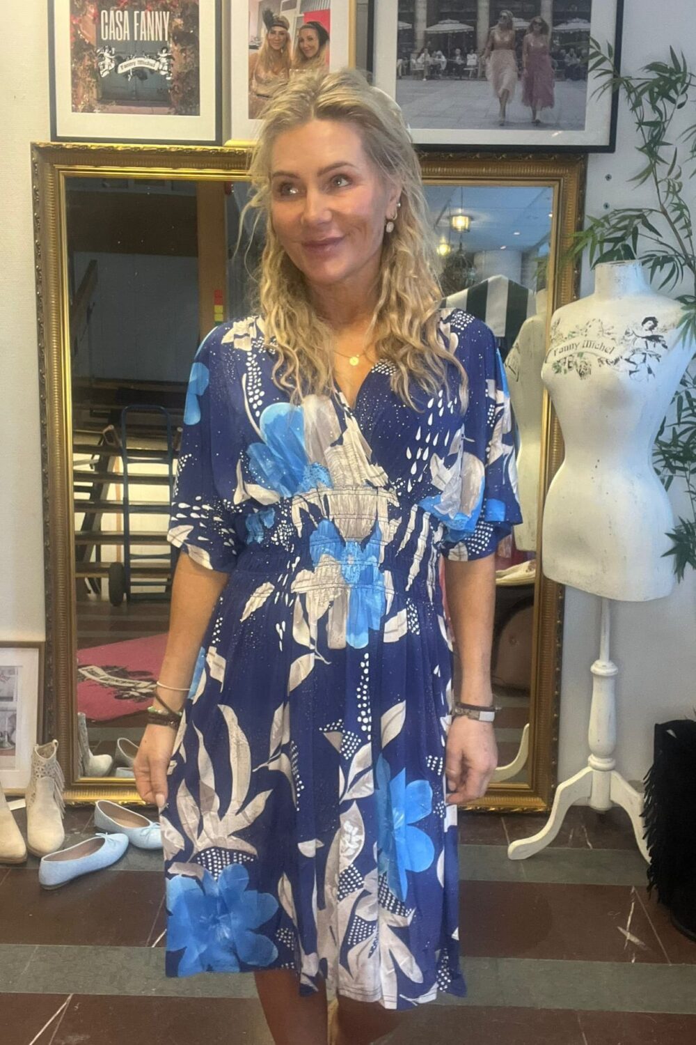 Kvinna i blå, blommig klänning står och ler lätt medan hon tittar åt sidan i en butik eller ateljé. Bakom henne syns en stor guldspegel, inramade foton och en provdocka med texten “Fanny Michel”.