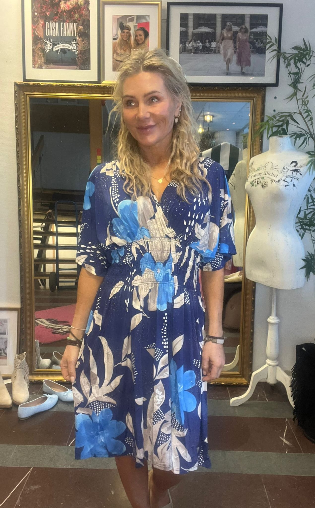 Kvinna i blå, blommig klänning står och ler lätt medan hon tittar åt sidan i en butik eller ateljé. Bakom henne syns en stor guldspegel, inramade foton och en provdocka med texten “Fanny Michel”.