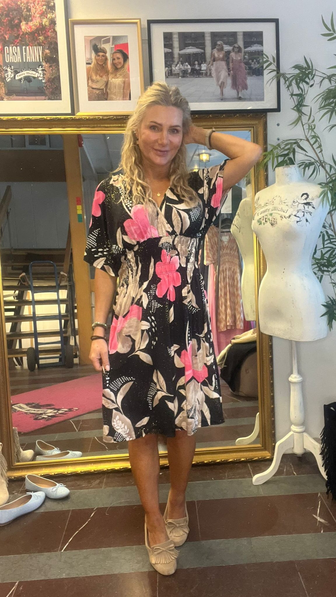 Kvinna står och poserar med ena handen bakom huvudet i en butik eller ateljé, klädd i svart klänning med stora rosa blommor och beige loafers. Bakom syns inramade foton och en provdocka. Text: “CASA FANNY”.