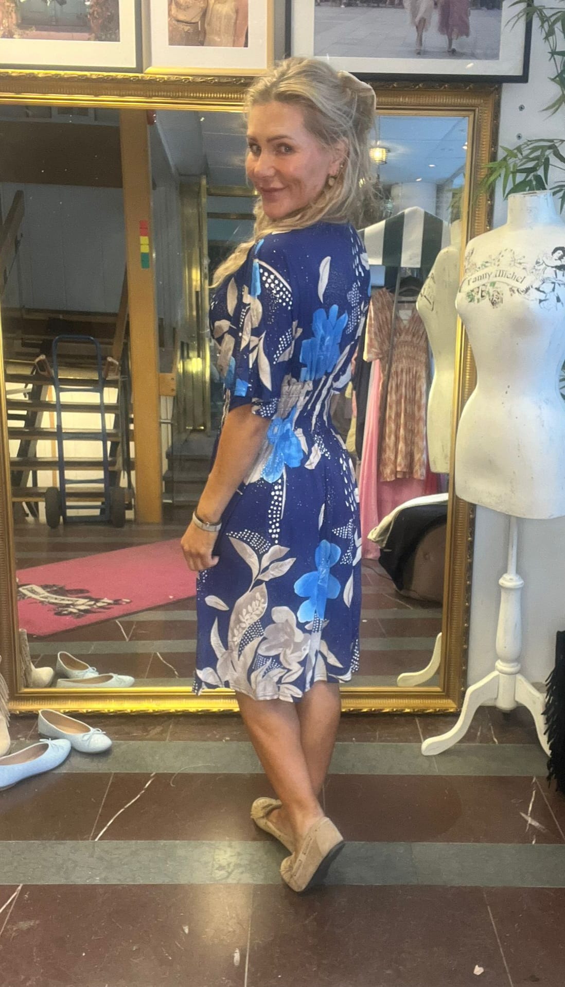 Blond kvinna står i profil och ler mot kameran medan hon provar en blå knälång klänning med stora blommönster. Hon står framför en spegel i en butik med skor på golvet och en provdocka bredvid.