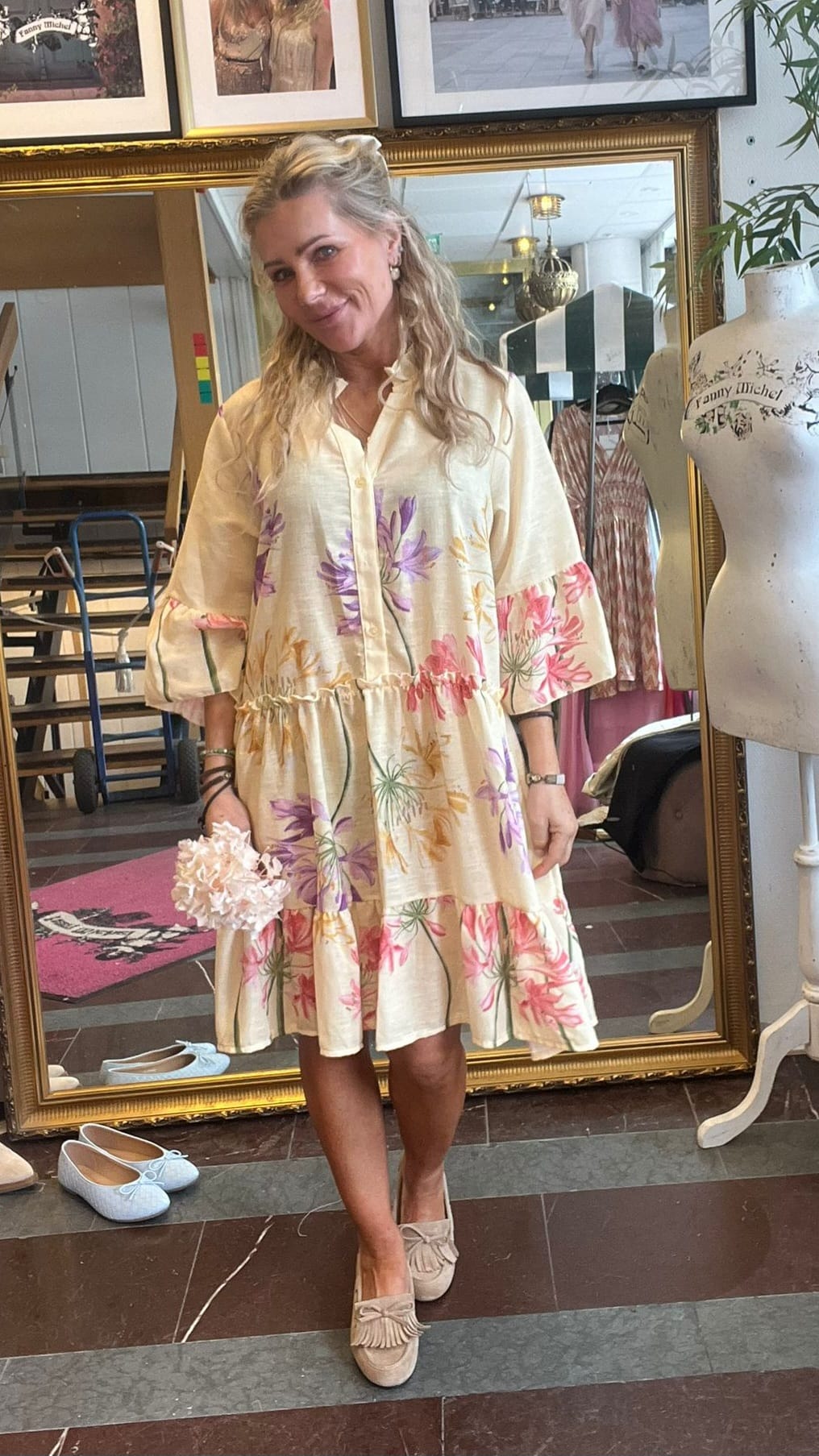 Blond kvinna står och poserar framför en stor guldramsspegel i en butik/ateljé, klädd i krämfärgad, blomsterprydd klänning och ljusa loafers. Runt henne syns provdocka med texten “Tammy Michel”, kläder och skor.