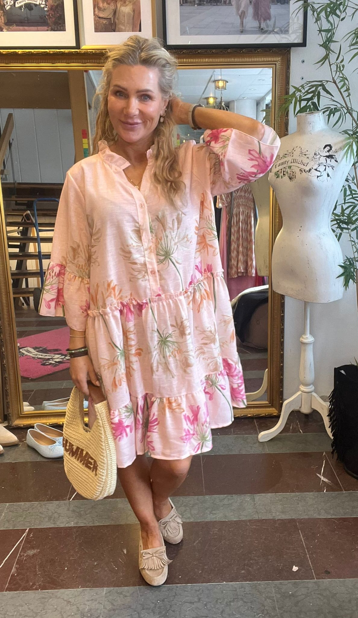 Leende kvinna poserar med ena handen bakom huvudet och håller en stråväska med texten ”CHANEL”. Hon bär en ljusrosa blommig klänning och loafers, i en butiksmiljö med speglar och provdocka märkt ”Fanny Michel”.