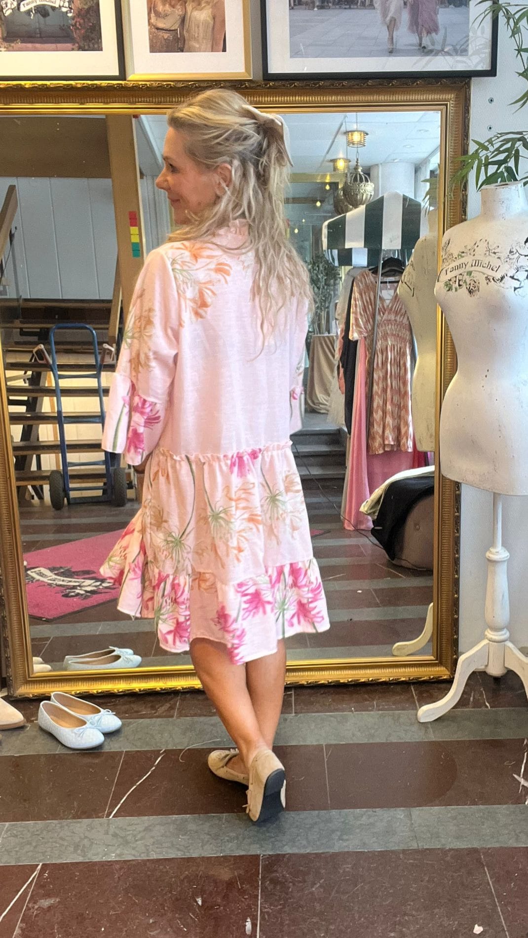 Kvinna i ljusrosa, blommig klänning står med ryggen mot kameran och tittar åt sidan framför en stor guldramsspegel i en butik. Bredvid syns en provdocka med texten ”Fanny Michel” och hängande klänningar.