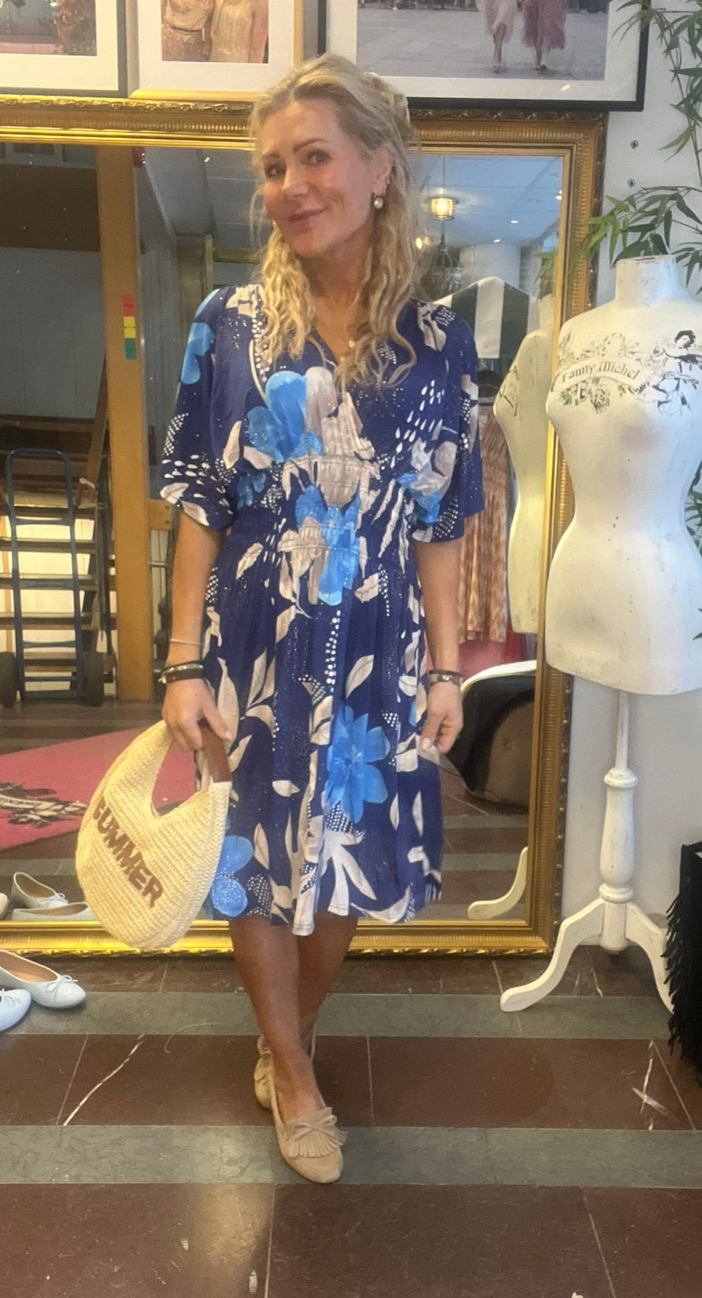 Kvinna står och ler framför en stor guldramad spegel, iklädd blå klänning med stora blommönster och ljusa loafers. Hon håller en halmväska med texten ”SUMMER”, i en butik med provdocka och skor.