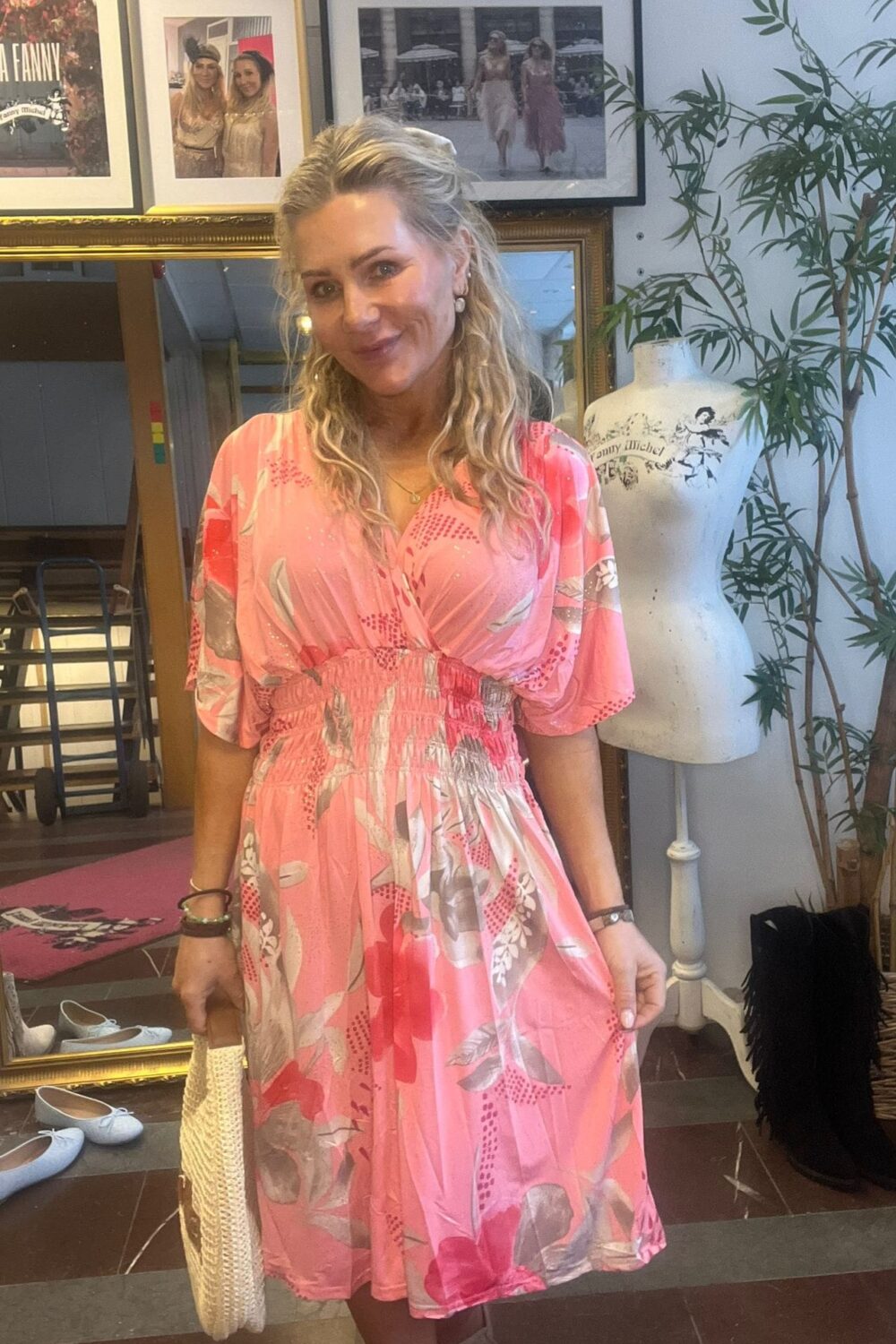Leende kvinna i rosa blommig klänning håller en ljus flätad väska och poserar i en butik/studio. I bakgrunden syns inramade foton, en spegel, en provdocka med texten “Fanny Michel” och en grön växt.