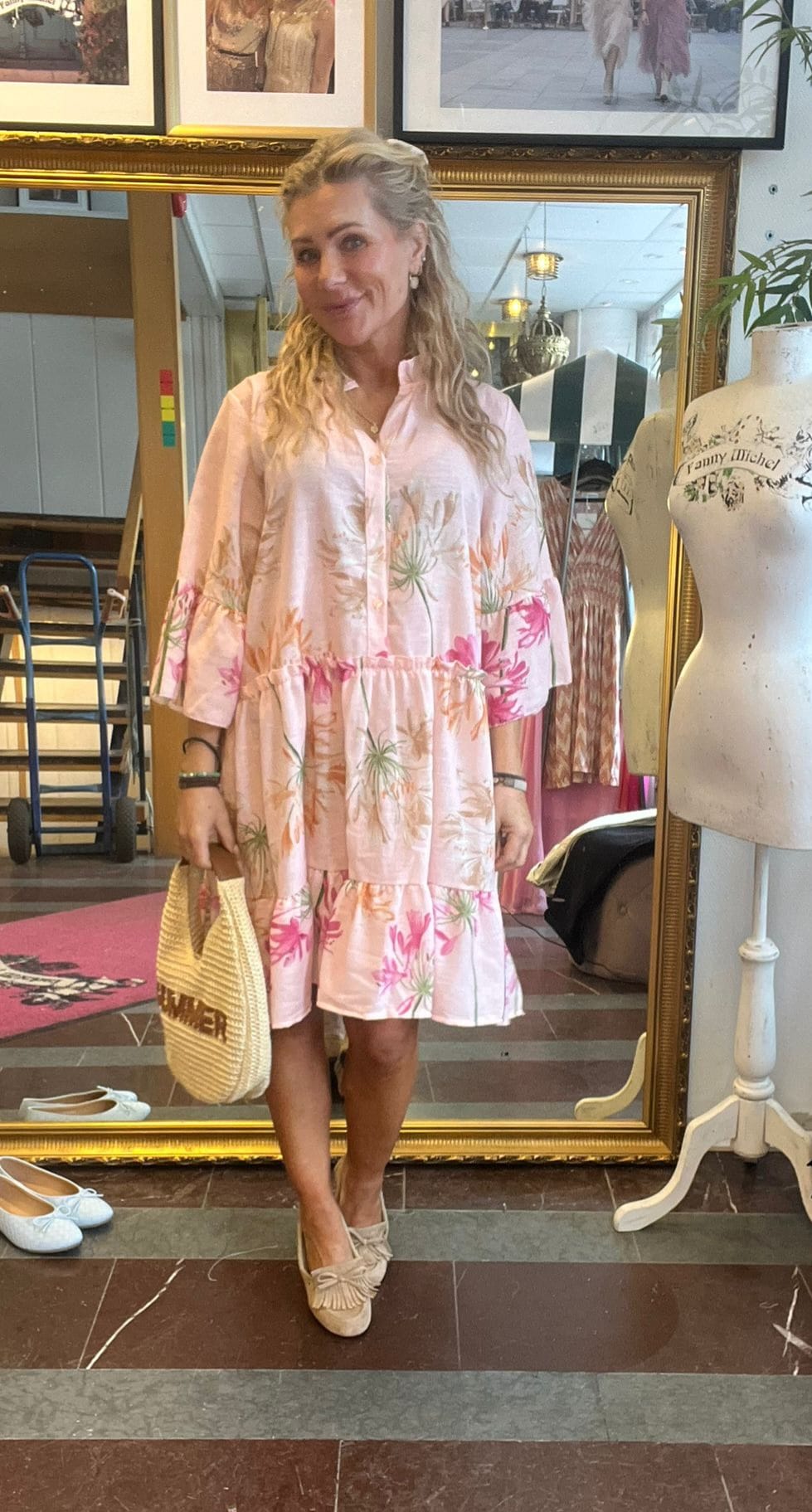Blond kvinna står och ler framför en stor guldramsspegel i en butik, iklädd ljusrosa blommig klänning och beige skor, och håller en stråväska. Intill syns skyltdocka med texten “Yanny Michel” och skor på golvet.