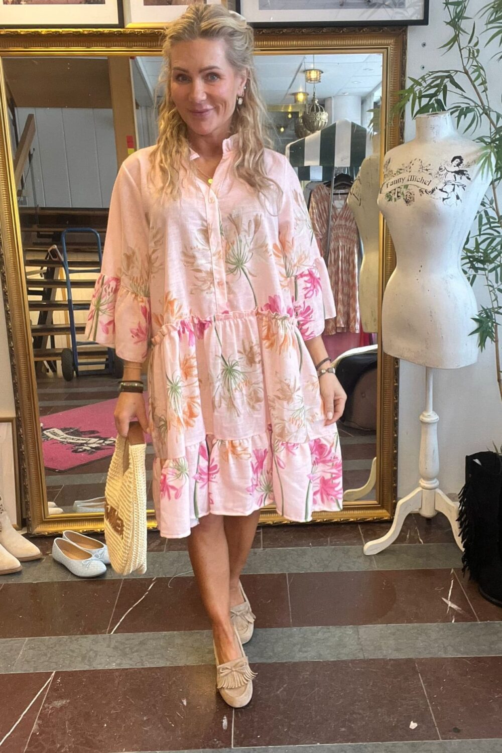 Leende kvinna står i en klädbutik och visar en ljusrosa, blommig volangklänning, med stråväska i handen och beige loafers. Bakom henne syns en stor guldramsspegel, klädställning och provdocka med texten ”Fanny Michel”.