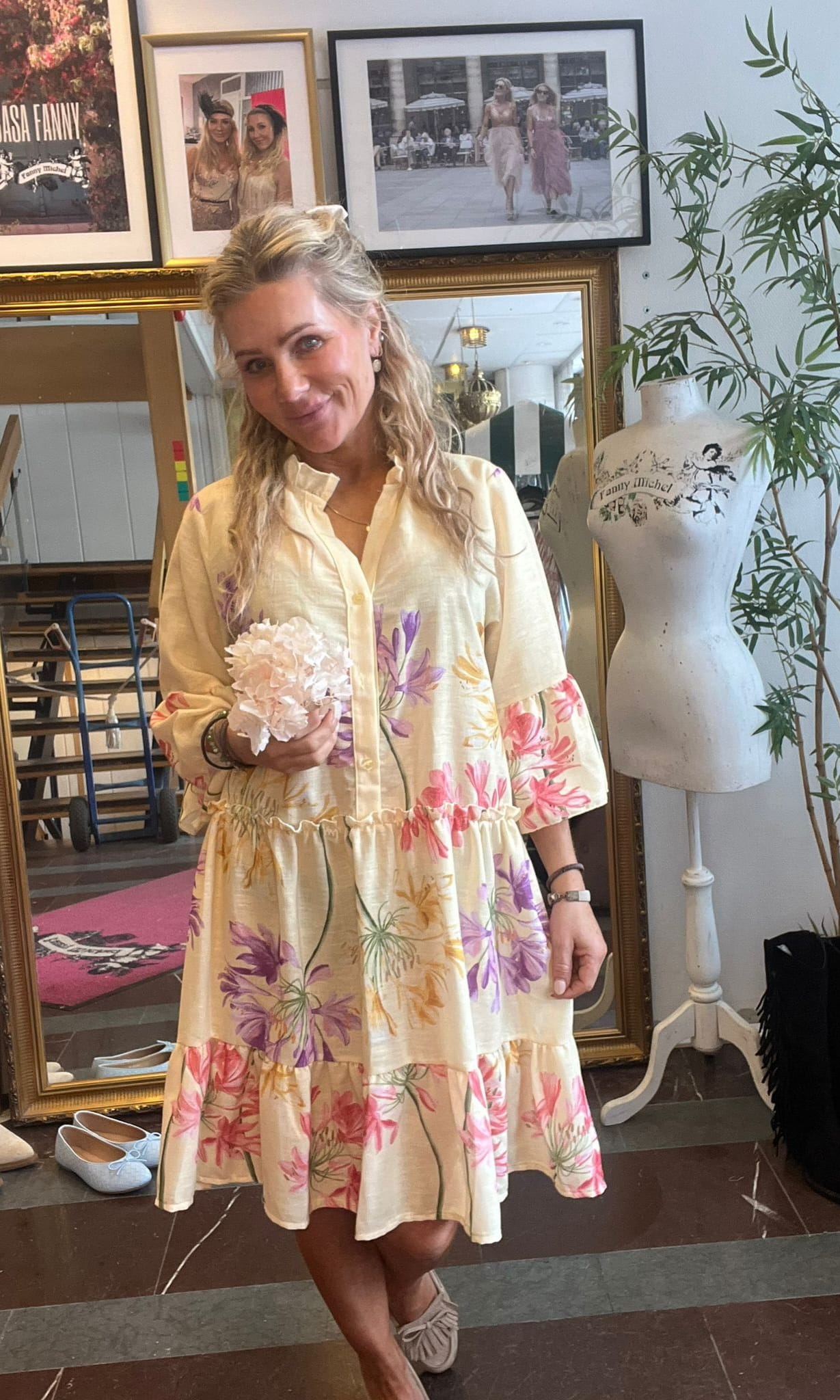 Leende kvinna i ljus blommig klänning håller en vit hortensia och står inomhus framför en spegel och inramade foton. Bredvid syns en provdocka med texten ”Fanny Michel” och en grön växt.