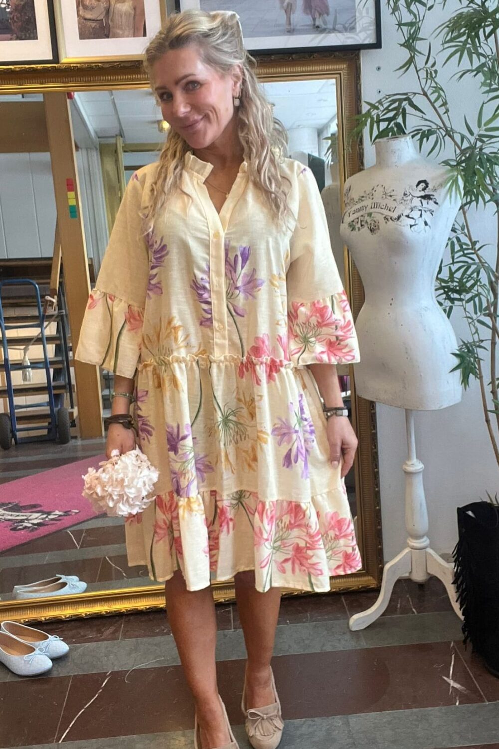 Blond kvinna står och ler i en butik/ateljé, iklädd en ljus klänning med stora blommönster och rosettprydda loafers, och håller ett litet blombukettlikt föremål. Bakom syns spegel, inramade foton och provdocka med texten “Fanny Michel”.
