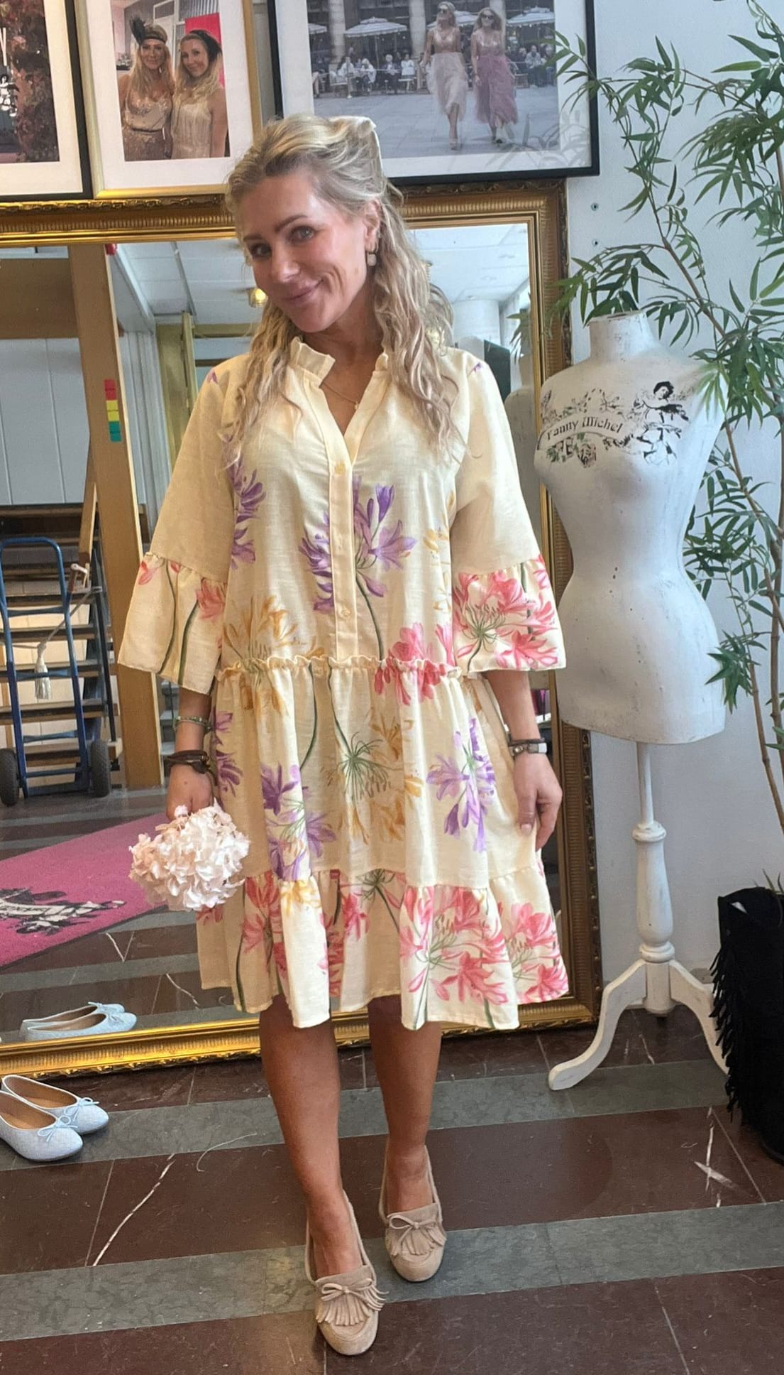 Blond kvinna står och ler i en butik/ateljé, iklädd en ljus klänning med stora blommönster och rosettprydda loafers, och håller ett litet blombukettlikt föremål. Bakom syns spegel, inramade foton och provdocka med texten “Fanny Michel”.