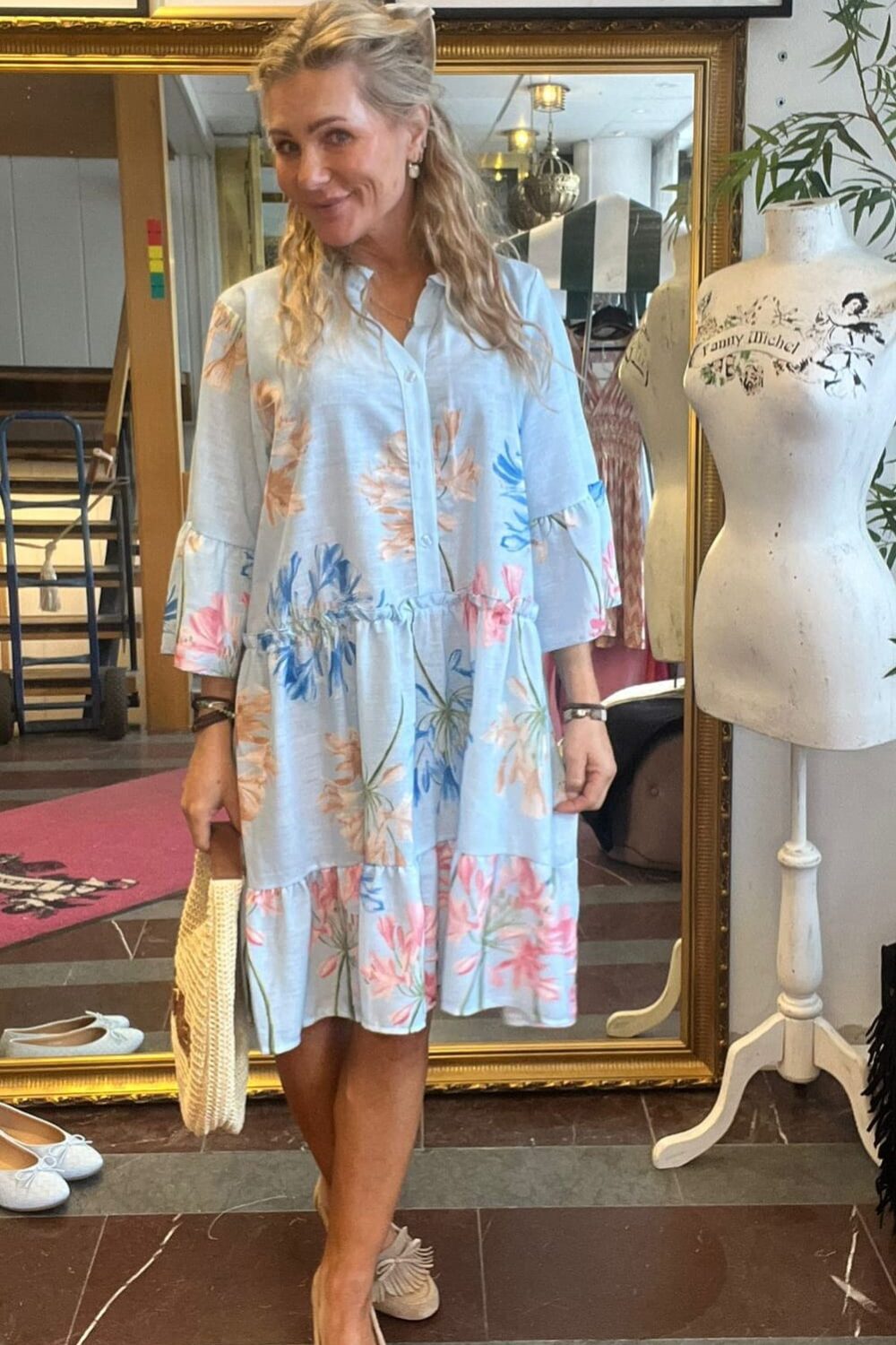 Leende kvinna står och poserar i en butik eller ateljé, klädd i ljusblå blommig klänning och beige skor, med stråväska i handen. Bakom henne syns stor guldramsspegel, foton på väggen och provdocka med texten ”Fanny Michel”.