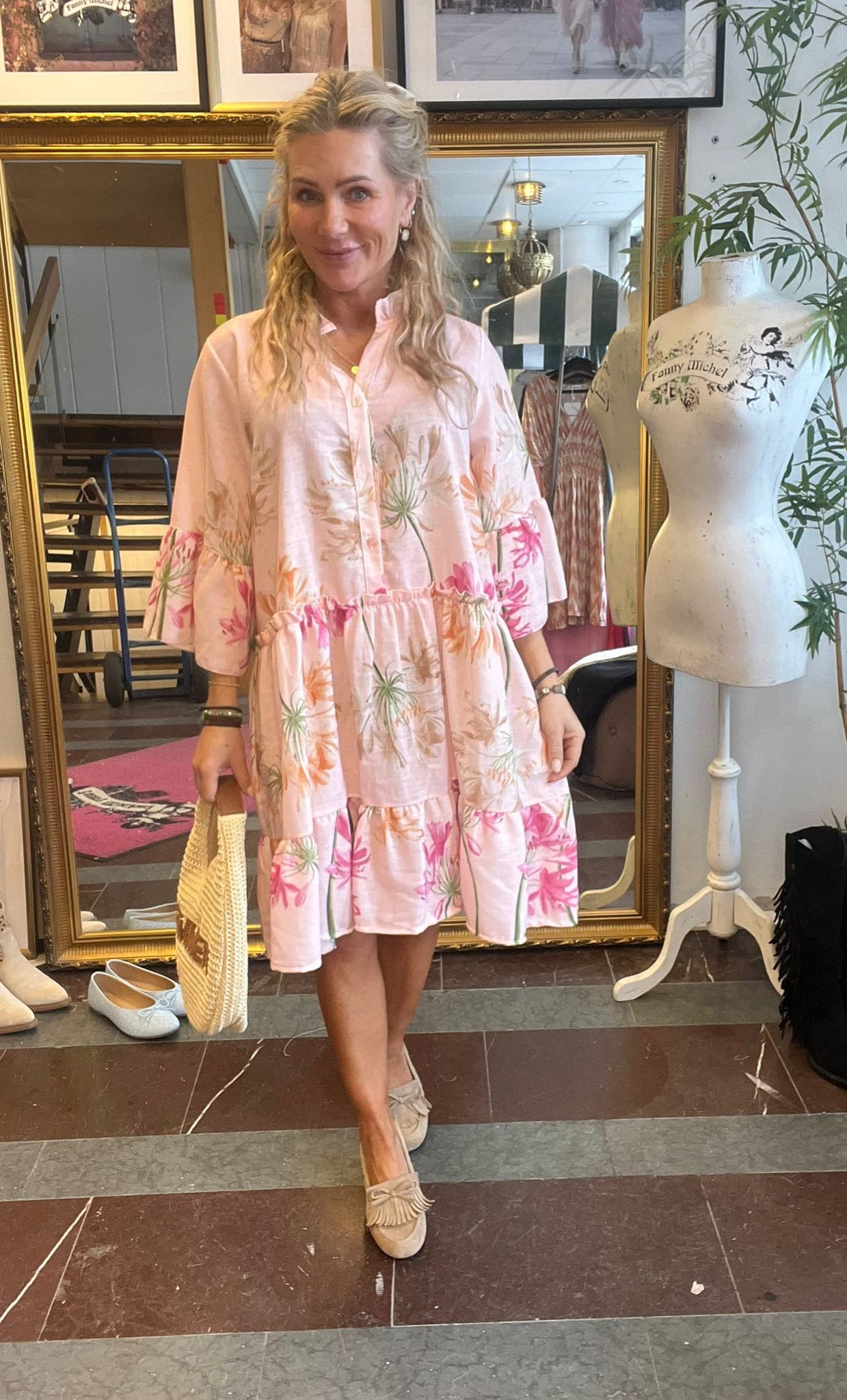 Leende kvinna står i en klädbutik och visar en ljusrosa, blommig volangklänning, med stråväska i handen och beige loafers. Bakom henne syns en stor guldramsspegel, klädställning och provdocka med texten ”Fanny Michel”.