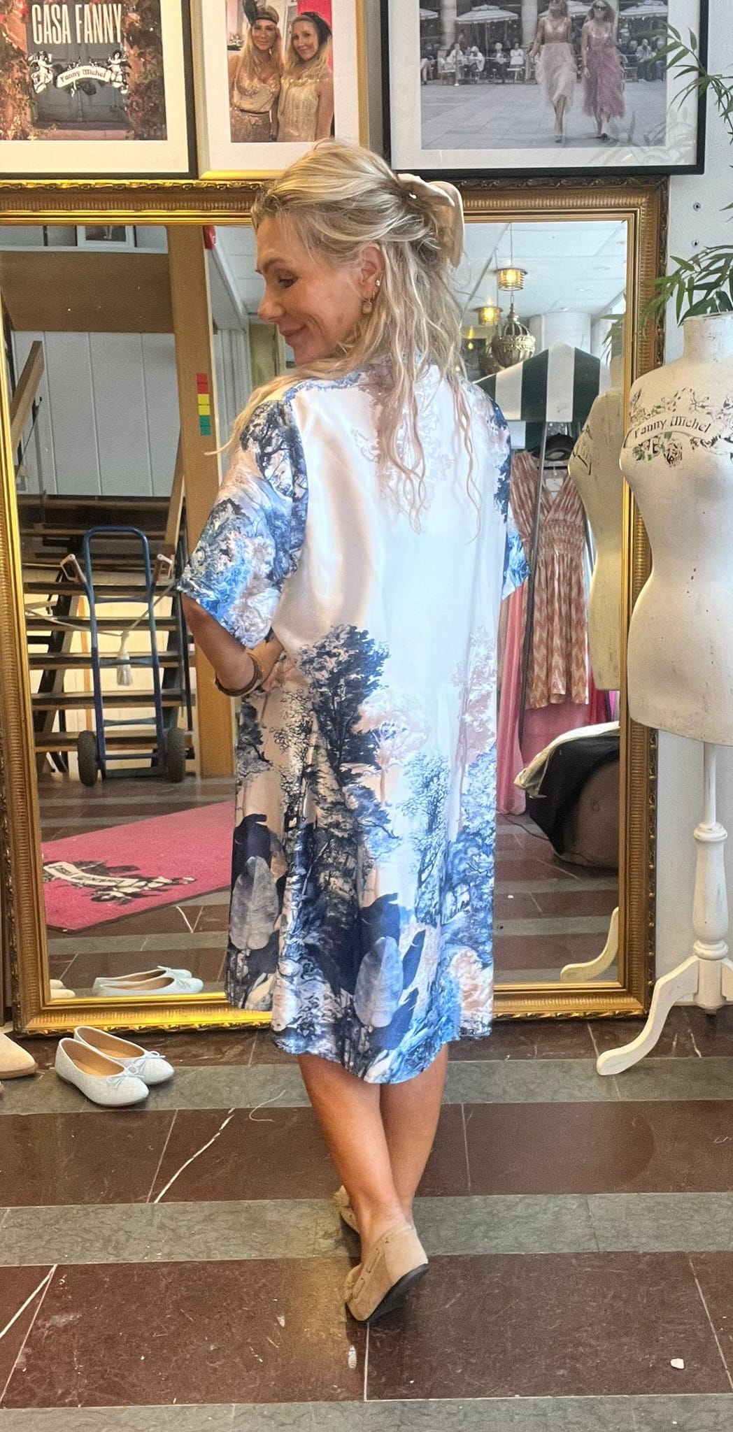 Blond kvinna står med ryggen mot kameran och ler åt sidan, medan hon visar upp en ljus klänning med blått landskapsmönster i en butik. Bakom syns guldramsspegel, skyltdocka med texten “Fanny Michel” och inramade foton.