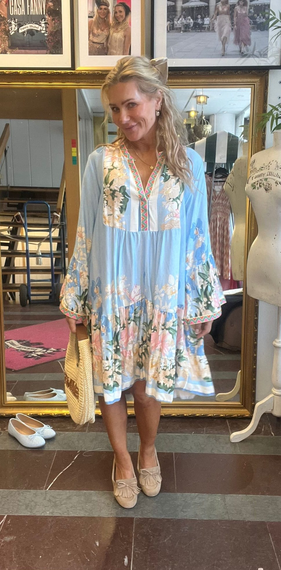 Blond kvinna står och ler framför en stor guldramad spegel, klädd i ljusblå blommig tunika med broderad kant, beige loafers och halmhatt i handen. I bakgrunden syns skylt “CASA FANNY”.