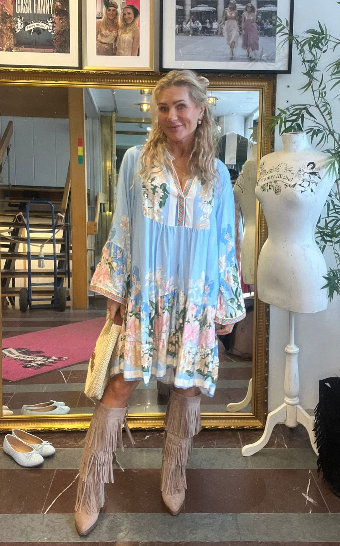 Leende kvinna poserar framför en stor spegel i en butik, klädd i ljusblå blommig tunika och fransstövlar samt håller en stråväska. Bredvid står en provdocka med texten “fanny michel”; inramade foton hänger ovanför.