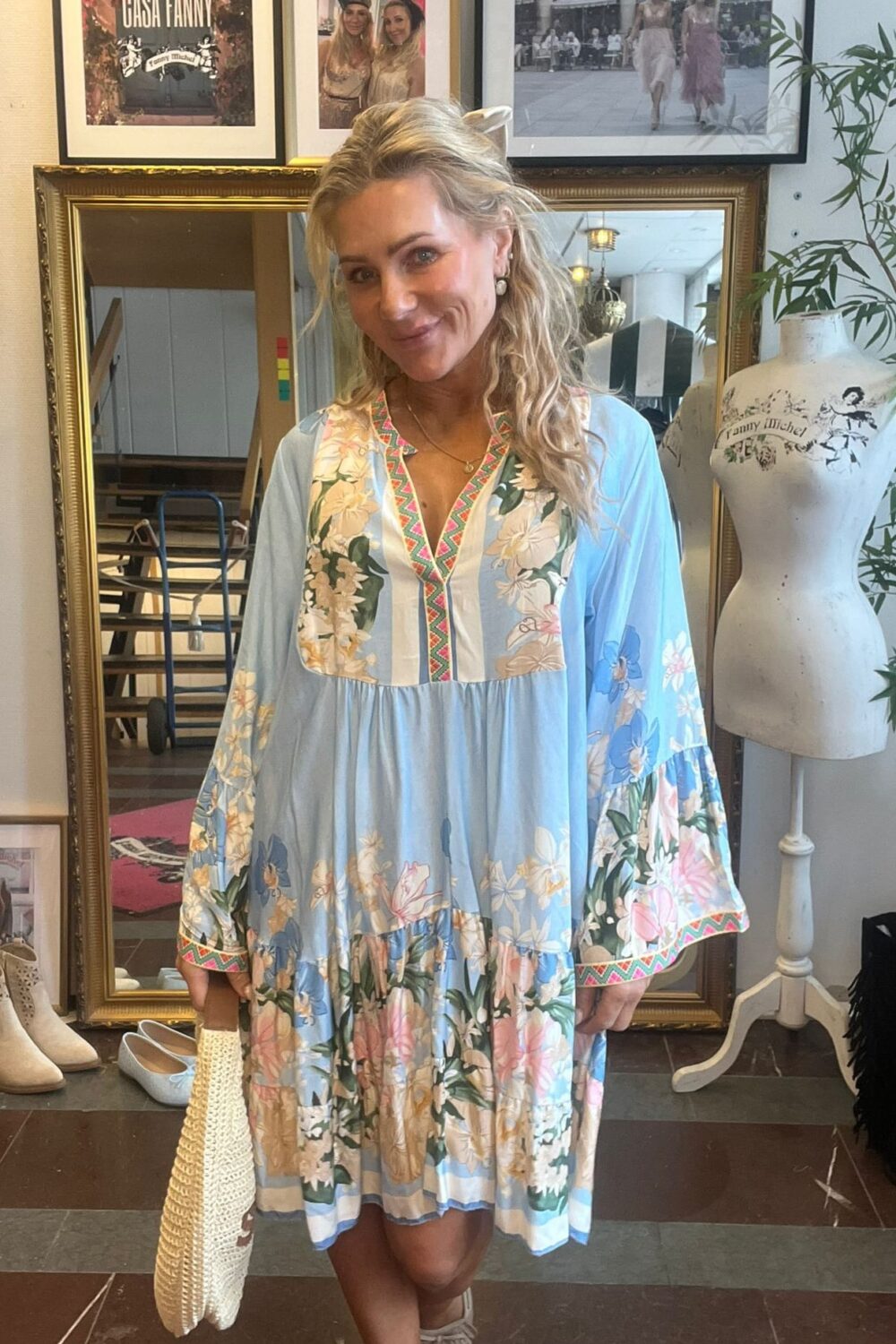 Blond kvinna ler mot kameran och står inomhus i en butik eller ateljé, klädd i ljusblå blommig klänning och håller en vävd väska. Bakom syns spegel, skylt “CASA FANNY” och provdocka med text “Fanny Michel”.