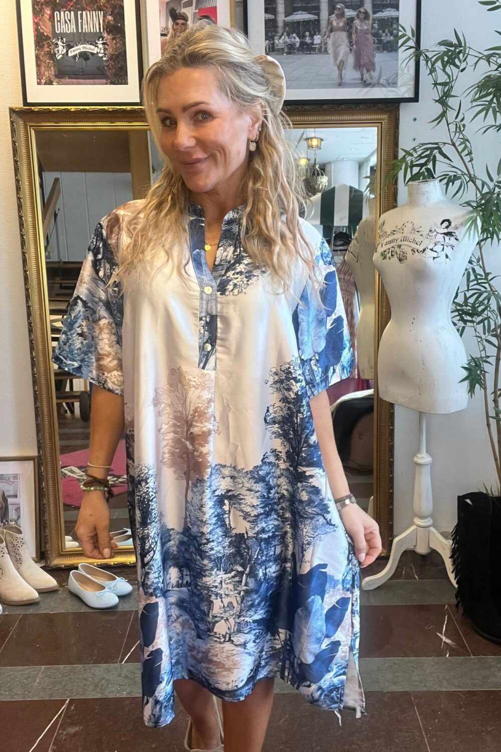 Blond kvinna ler och håller ut kjolen på en luftig blåvit mönstrad skjortklänning i en butik/ateljé. Bakom syns guldspegel, skor på golvet, provdocka med texten ”Fanny Michel” och inramad affisch ”CASA FANNY”.