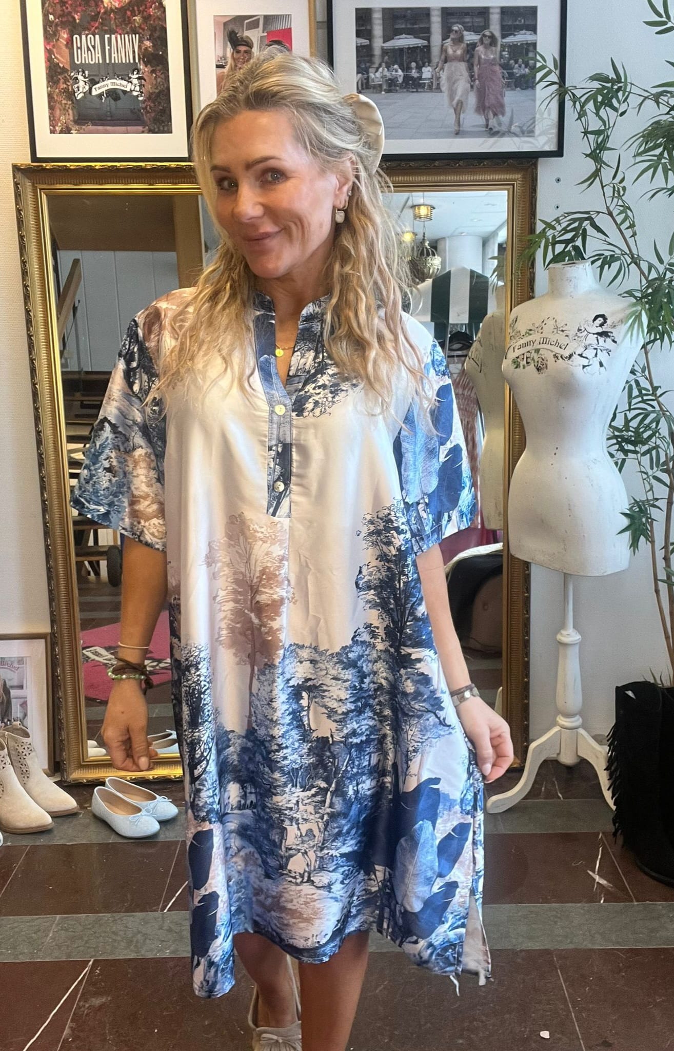 Blond kvinna ler och håller ut kjolen på en luftig blåvit mönstrad skjortklänning i en butik/ateljé. Bakom syns guldspegel, skor på golvet, provdocka med texten ”Fanny Michel” och inramad affisch ”CASA FANNY”.