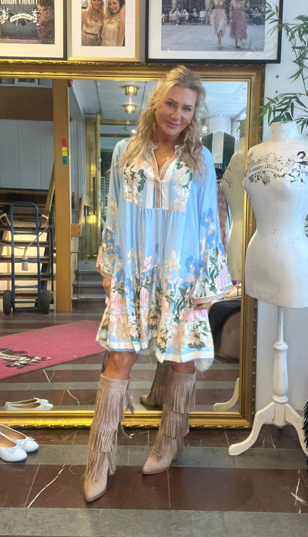 Kvinna poserar och ler framför en stor spegel i en butik eller ateljé, klädd i ljusblå blommig klänning och beige fransstövlar. Bredvid står en provdocka med texten “Tamy Michel” och inramade foton hänger ovanför.