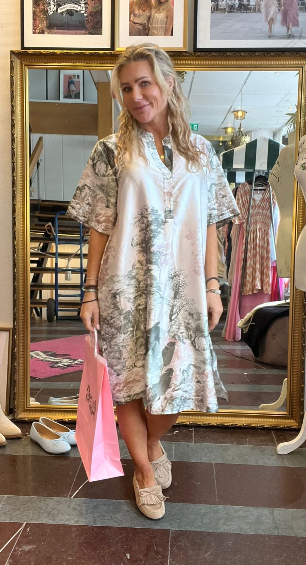 Blond kvinna står framför en stor guldramsspegel och poserar med en rosa shoppingpåse. Hon bär en luftig, mönstrad skjortklänning och ljusa loafers; bakom syns klädställningar och inredning i en butik.