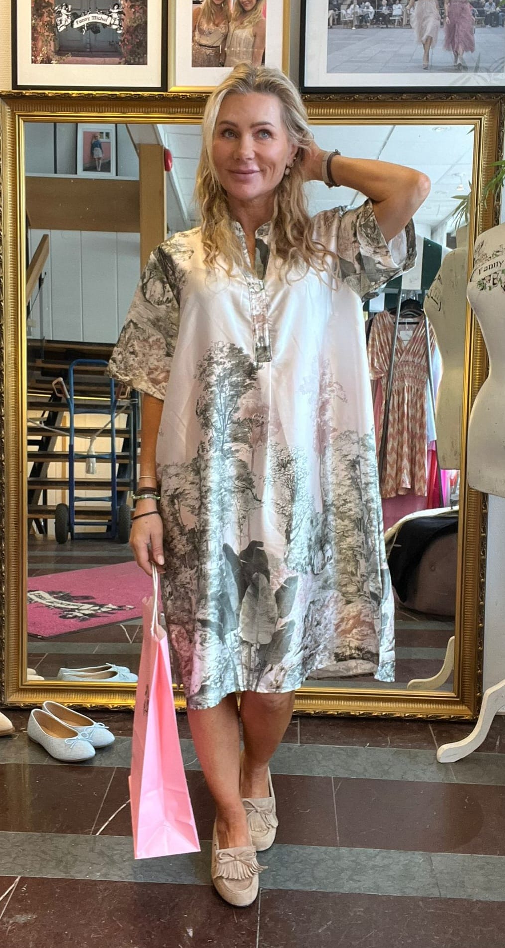 Blond kvinna står och poserar framför en stor guldramad spegel, håller en rosa shoppingpåse och har ena handen i håret. Hon bär en lös mönstrad klänning och ljusa loafers i en butiksmiljö med skor och ramar.