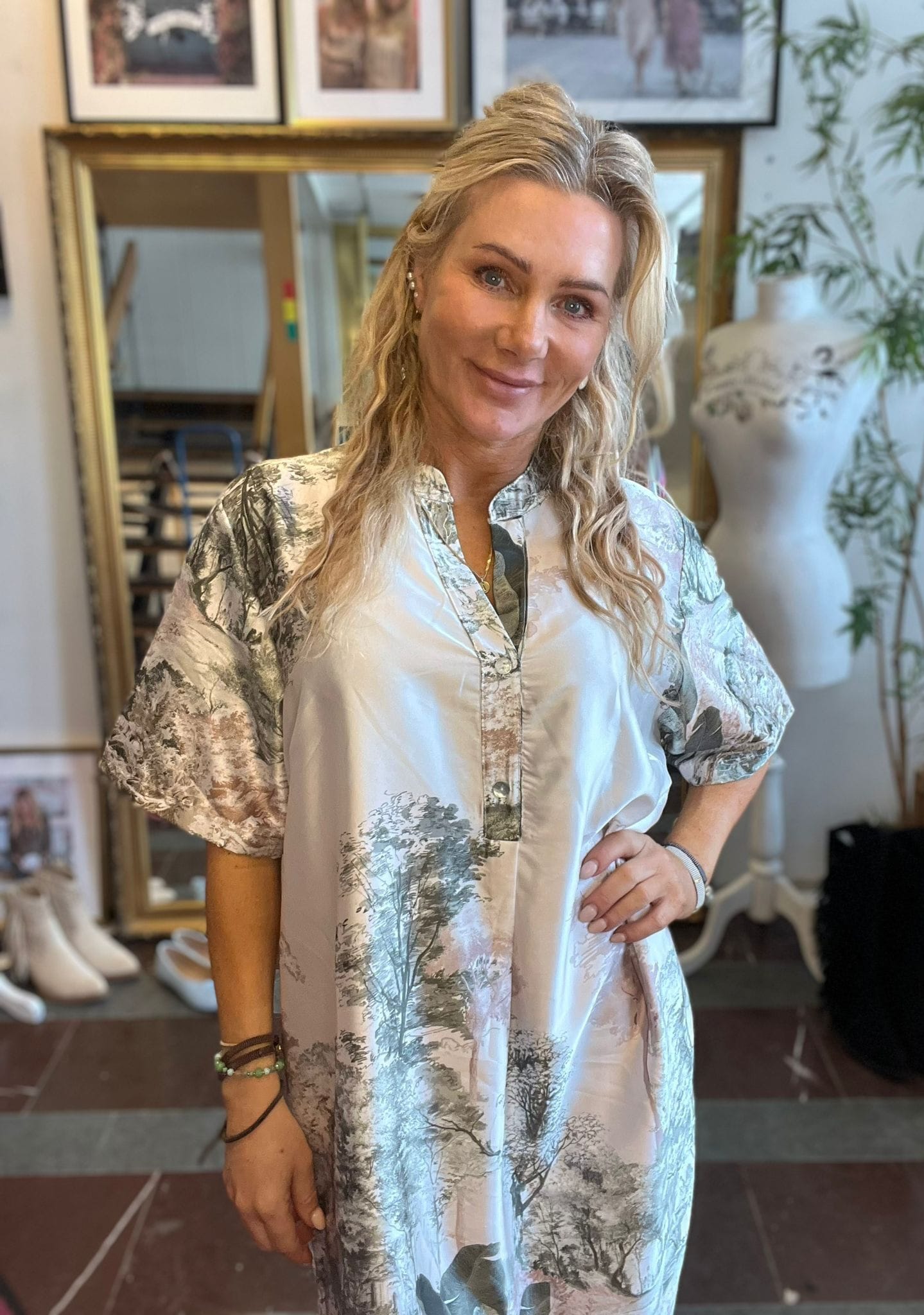 Leende kvinna med långt blont hår poserar med ena handen på höften och bär en ljus, mönstrad klänning. Hon står inomhus framför en stor spegel, inramade foton, en provdocka och vita skor.