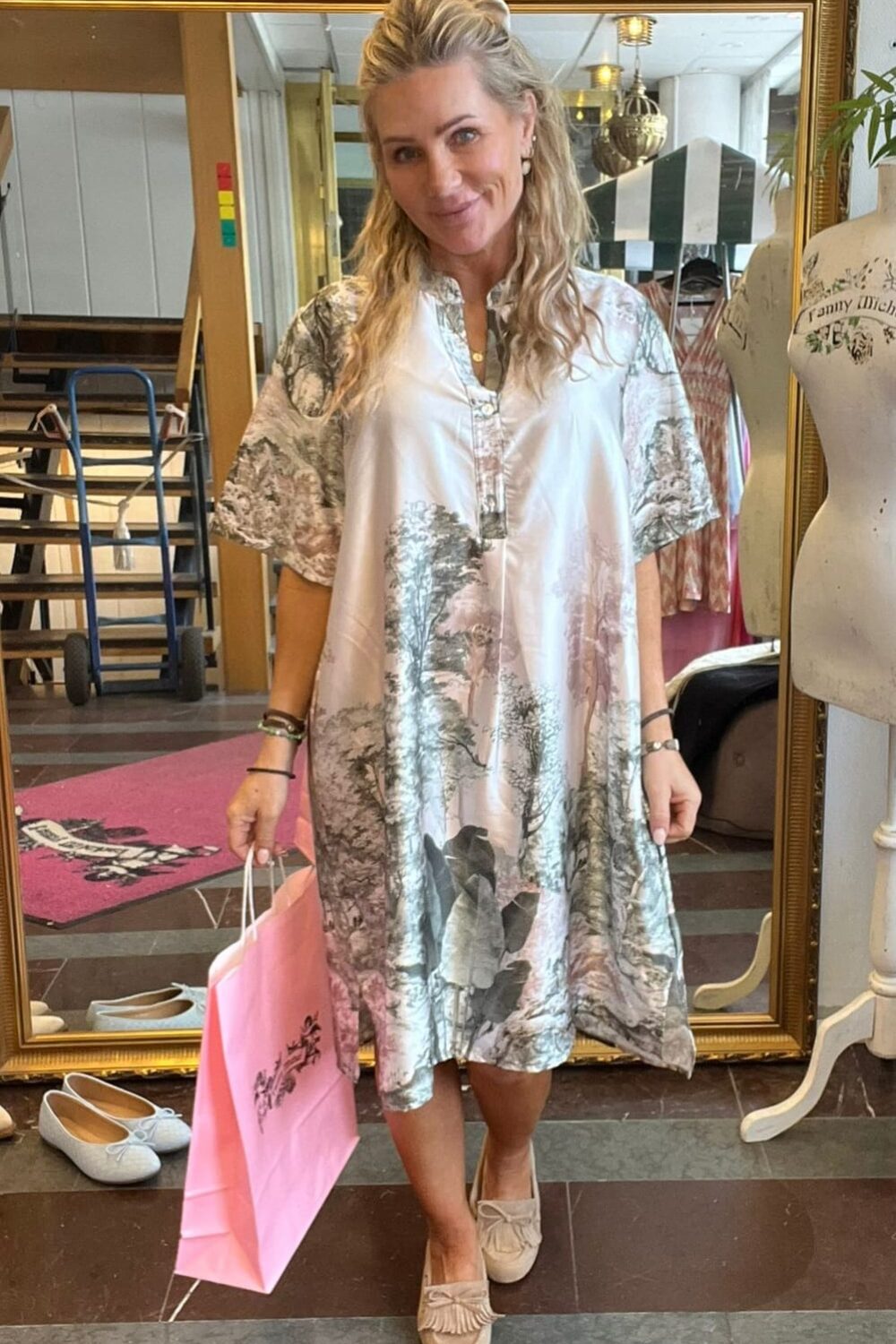 Blond kvinna står och ler i en klädbutik, håller en rosa shoppingpåse och visar en luftig, mönstrad klänning med kort ärm. I bakgrunden syns en stor guldramsspegel, skor på golvet och en provdocka.