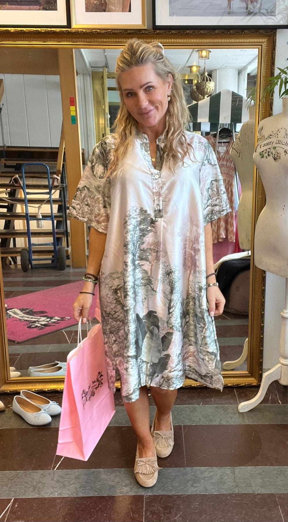 Blond kvinna står och ler i en klädbutik, håller en rosa shoppingpåse och visar en luftig, mönstrad klänning med kort ärm. I bakgrunden syns en stor guldramsspegel, skor på golvet och en provdocka.