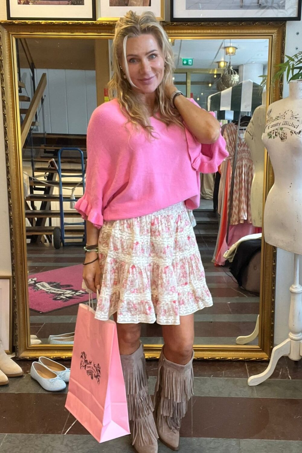Leende kvinna poserar i en klädbutik framför en stor guldramsspegel, med handen vid håret och en rosa shoppingpåse. Hon bär rosa tröja, mönstrad volangkjol och fransstövlar; skylten “CASA FANNY” syns ovanför.