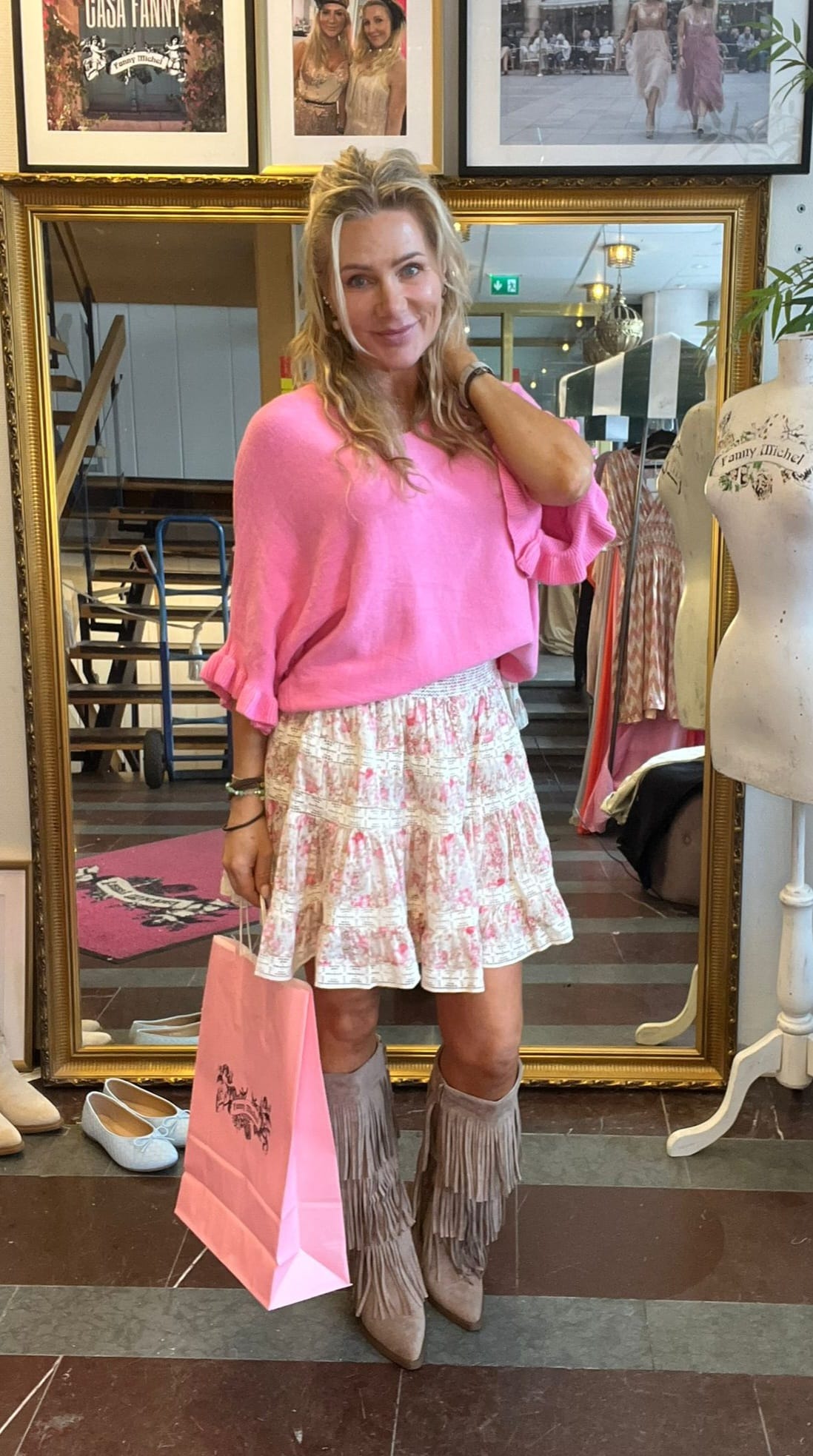 Leende kvinna poserar i en klädbutik framför en stor guldramsspegel, med handen vid håret och en rosa shoppingpåse. Hon bär rosa tröja, mönstrad volangkjol och fransstövlar; skylten “CASA FANNY” syns ovanför.