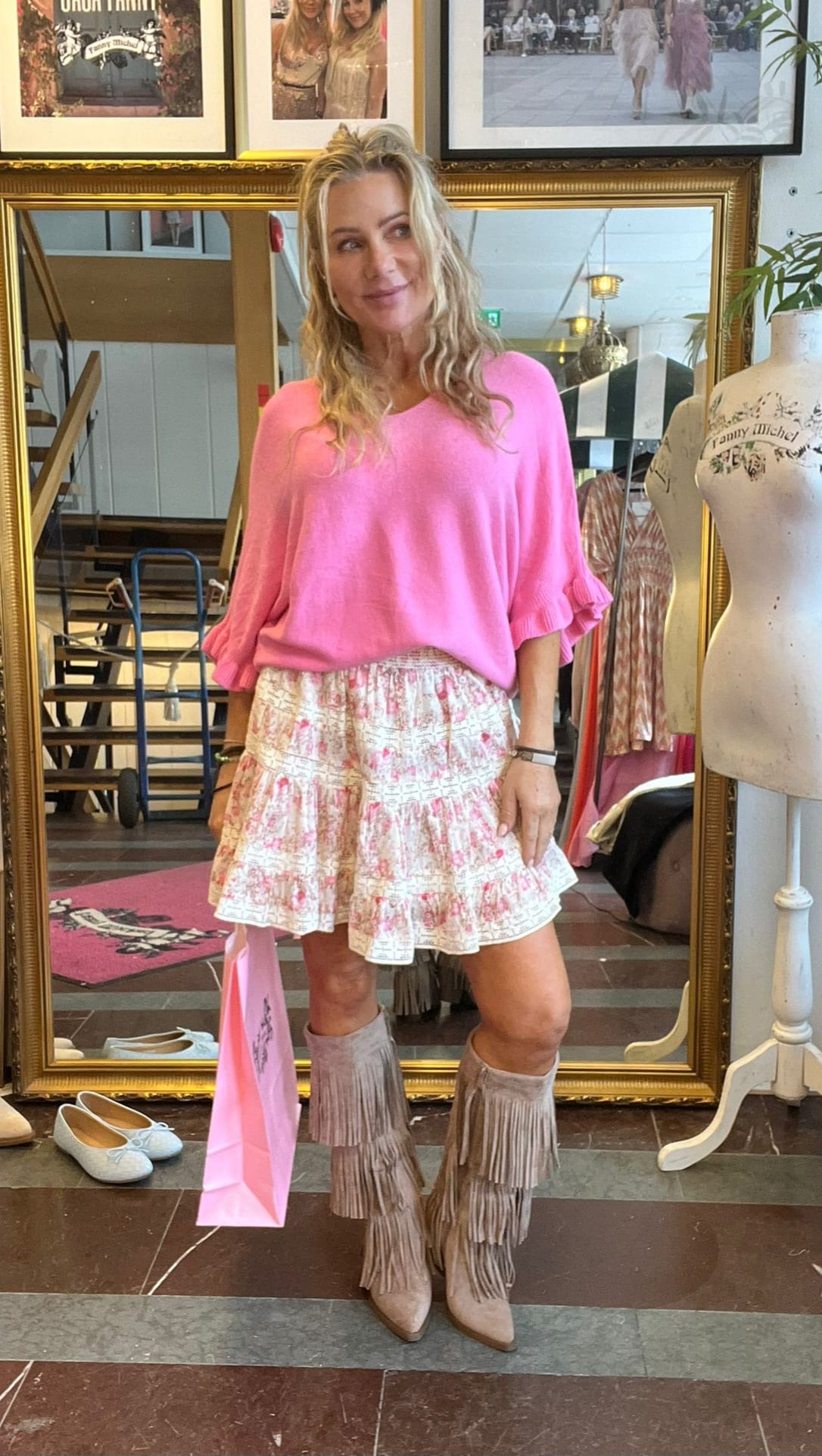 Leende kvinna poserar framför en stor guldramsspegel i en klädbutik, iklädd rosa tröja, blommig volangkjol och fransstövlar. Hon håller en rosa shoppingpåse; bakom syns klädställning, trappa och skyltdocka.