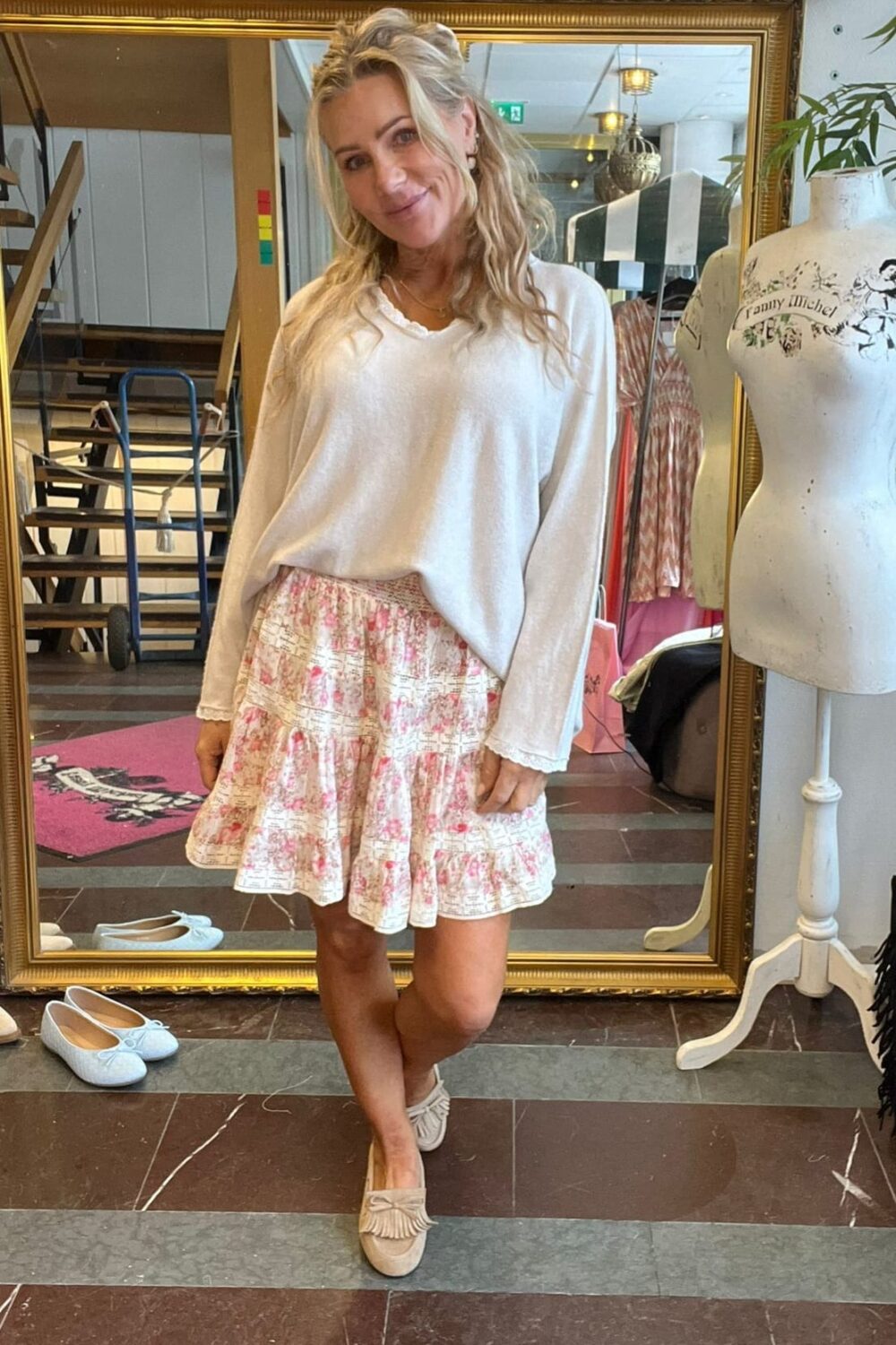 Blond kvinna ler och poserar framför en stor guldramsspegel i en klädbutik, klädd i vit tröja och rosa blommig volangkjol med beige loafers. Bredvid står en provdocka med texten “Fanny Michel”.