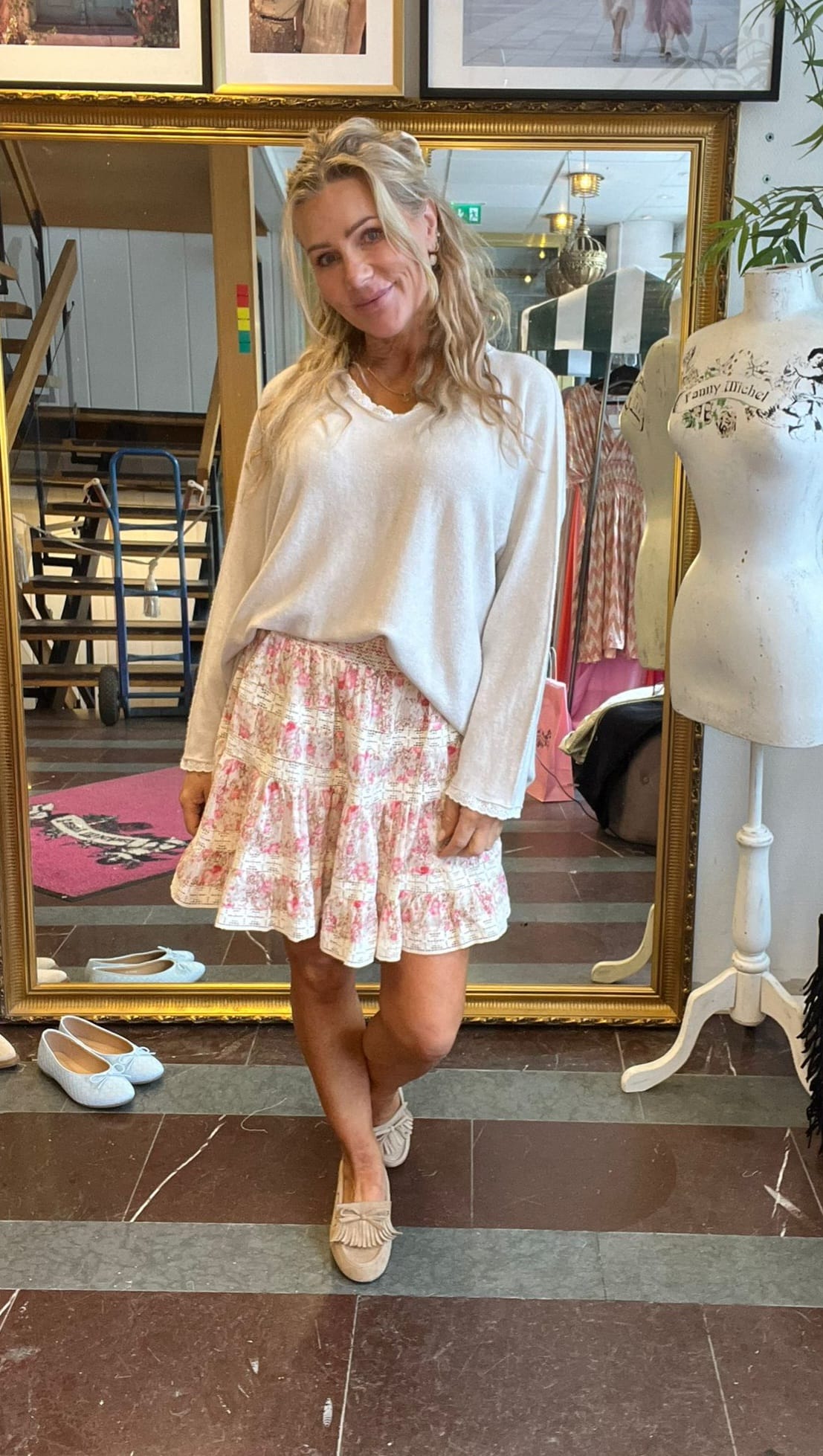 Blond kvinna poserar och ler framför en stor guldramsspegel i en butik. Hon bär vit långärmad tröja, blommig volangkjol och beige loafers. Intill syns en provdocka med texten “Fanny Michel”.