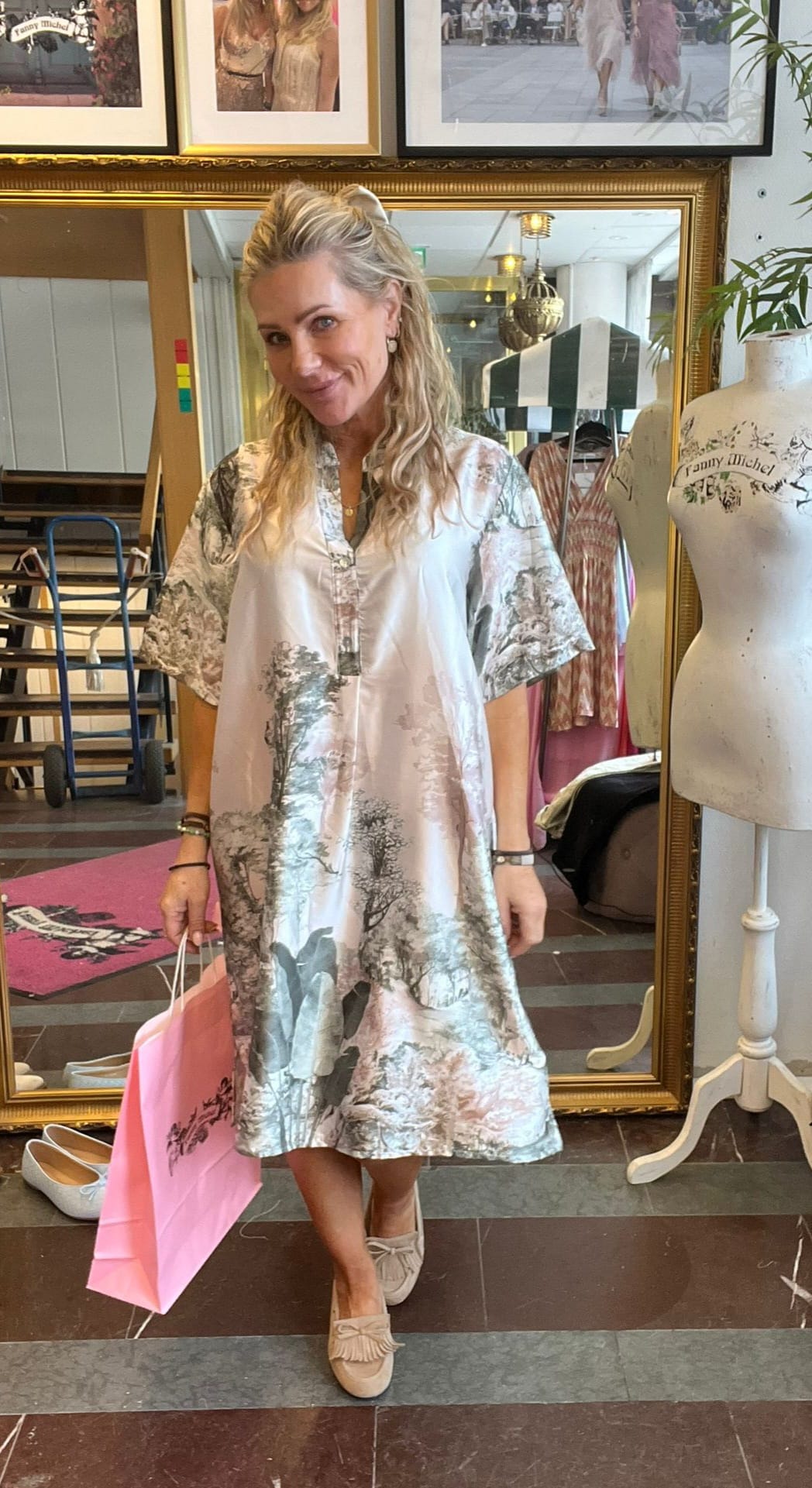 Blond kvinna poserar och ler i en klädbutik, håller en rosa shoppingpåse. Hon bär en luftig, ljus klänning med naturmotiv och beige skor. Bakom syns stor spegel, klänningar och provdocka med texten ”Tommy Michel”.