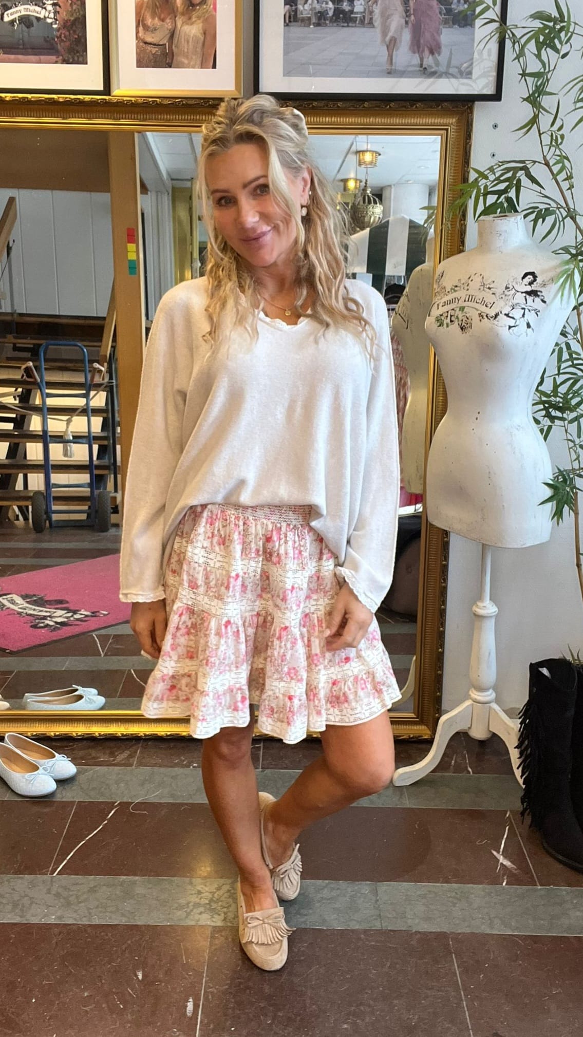 Blond kvinna poserar och ler i en butik eller ateljé, klädd i vit långärmad tröja, blommig volangkjol och ljusa loafers. Bredvid står en provdocka med texten ”Fanny Michel”, och speglar samt skor syns i bakgrunden.