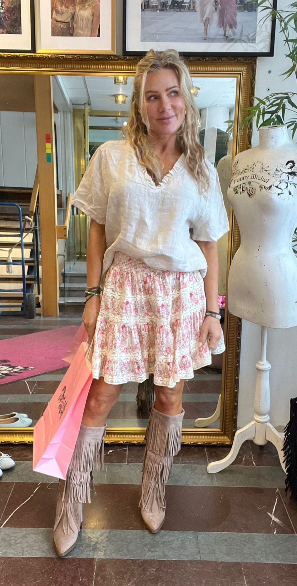 Blond kvinna står och ler framför en stor guldramsspegel, håller en rosa shoppingpåse och bär vit blus, blommig volangkjol och fransstövlar. Miljön liknar en butik med provdocka och inramade foton.