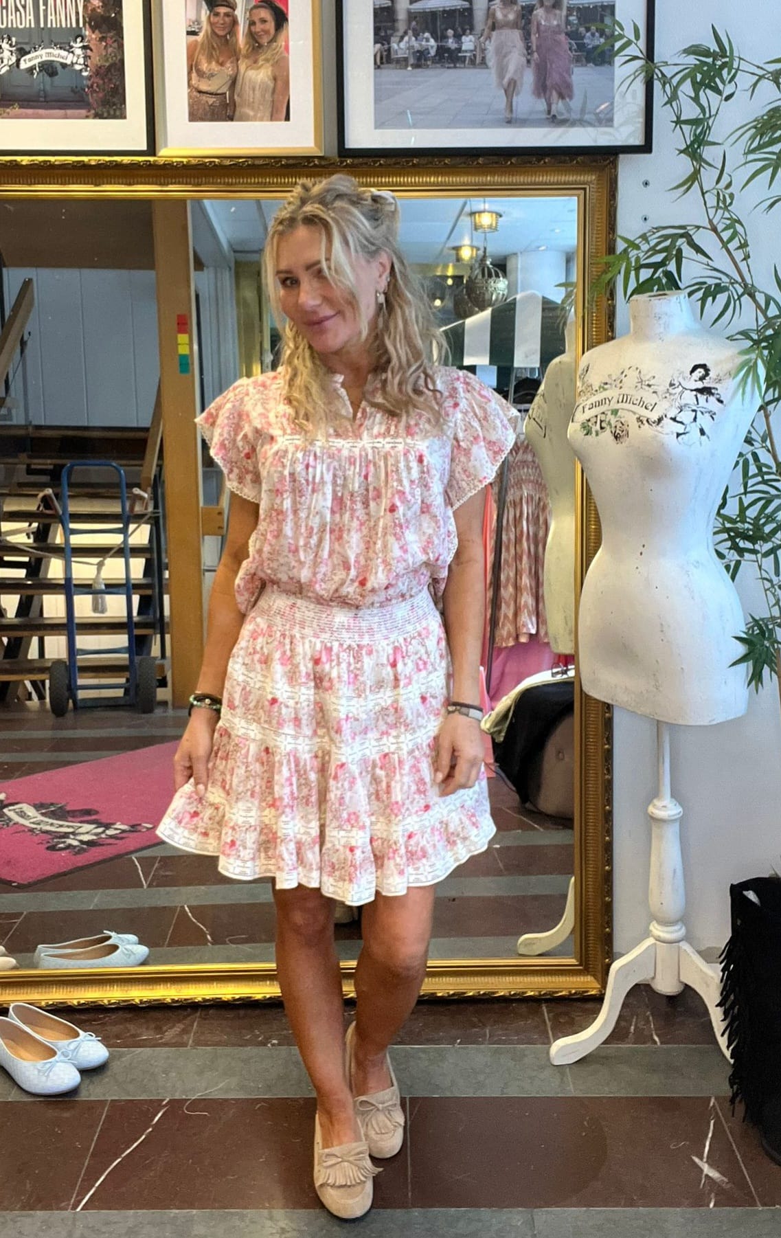 Kvinna står och poserar framför en stor guldramsspegel i en butik, klädd i kort rosa‑vit blommig klänning och beige loafers. Bredvid syns en provdocka med texten “Fanny Michel” och inramade foton på väggen.