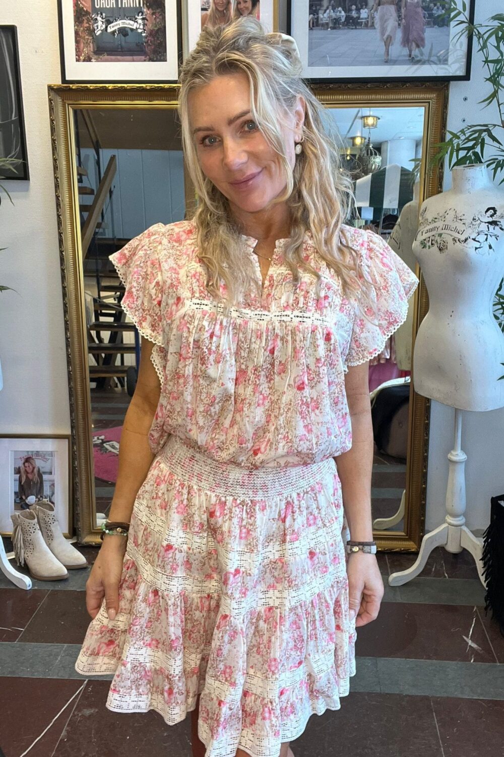 Leende kvinna med vågigt blont hår står och poserar i en rosa- och vitblommig klänning. Hon befinner sig i en butik eller studio med stor guldramsspegel, skyltdocka och inramade foton, varav ett har texten “Tanny Michel”.