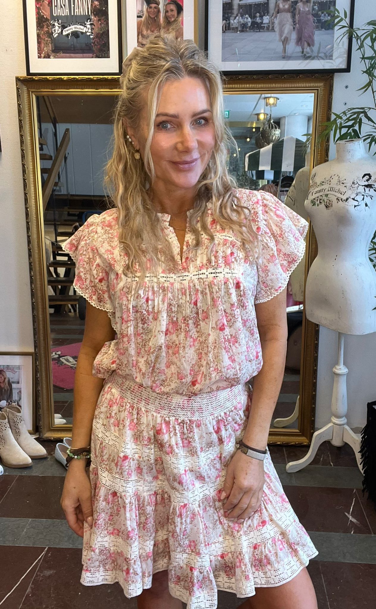 Leende kvinna med halvuppsatt blont hår poserar i en rosa- och vitblommig klänning inomhus. Bakom henne syns guldramade speglar och inramade bilder, samt en provdocka med texten ”Fanny Michel”.