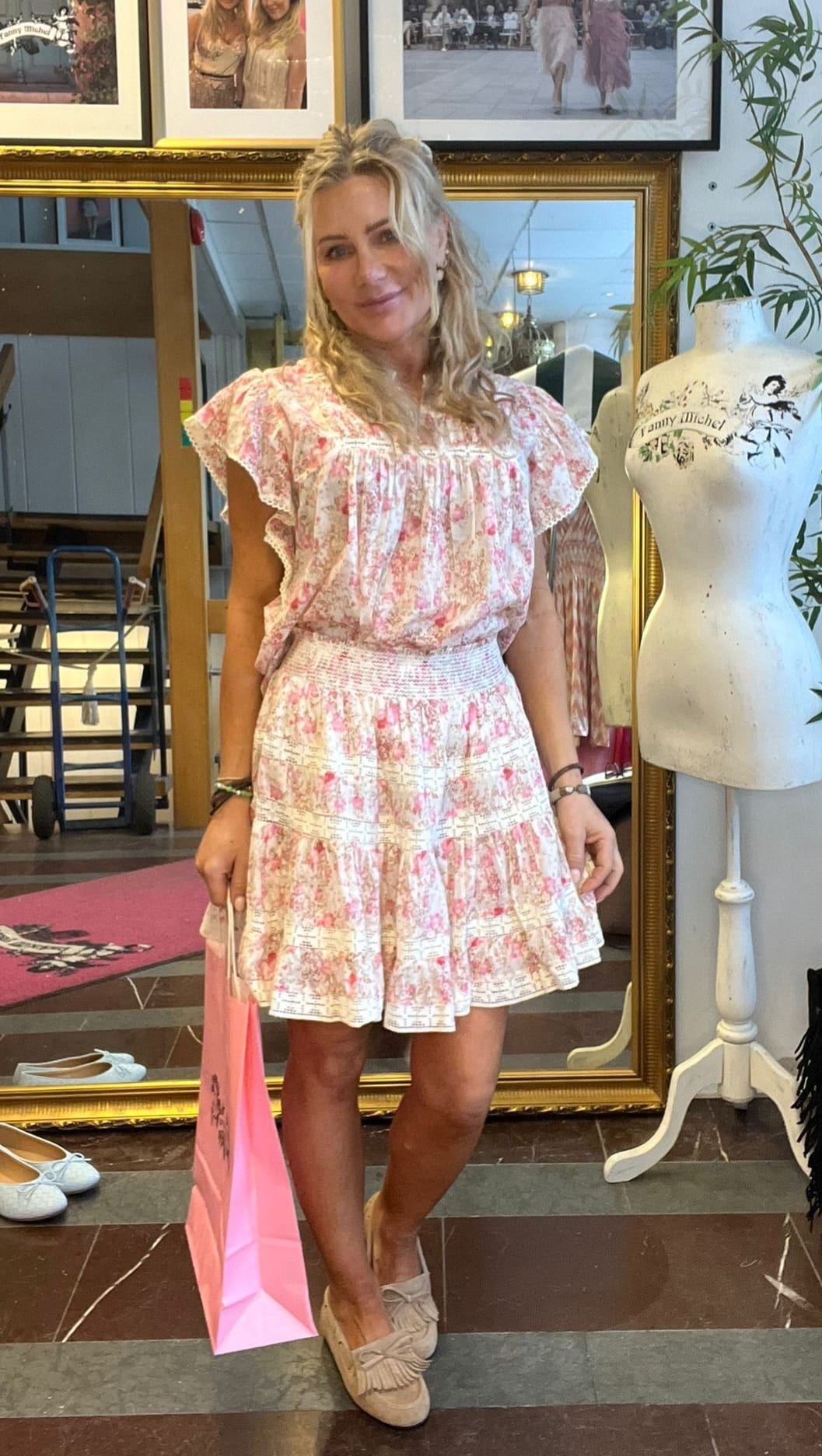 Blond kvinna står och ler i en butik, klädd i en ljusrosa blommig klänning och beige loafers, med en rosa shoppingpåse i handen. Bakom henne syns en stor spegel, inramade foton och en provdocka med texten “Jenny Michel”.