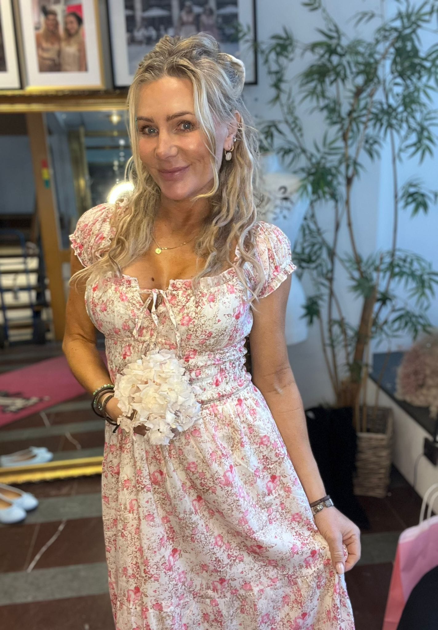 Blond kvinna i ljus, rosa blommig klänning poserar och ler mot kameran, med en vit blomsterbukett i handen. Hon står i ett rum med inramade foton, spegel och stor grön växt i bakgrunden.