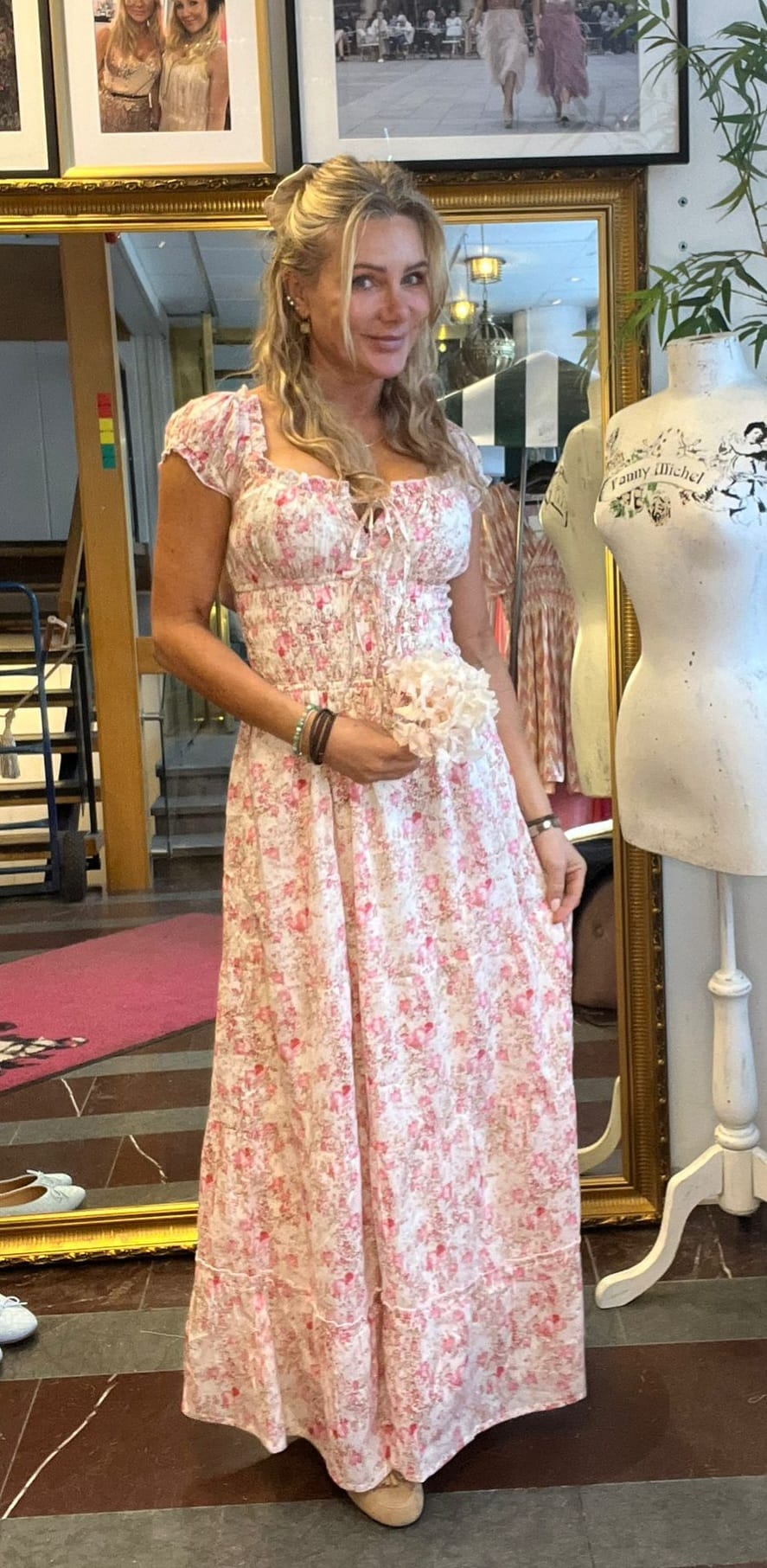 Blond kvinna står och poserar med en ljusrosa blommig långklänning och håller en liten blomsterbukett. Hon är i en ateljé/butik framför stor spegel, intill en provdocka med texten “Fanny Michel”.