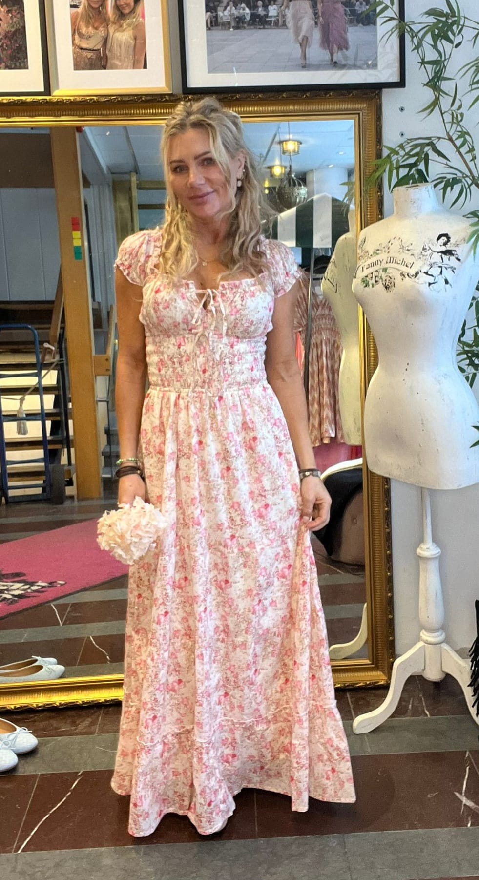 Blond kvinna står och ler i en butikslik miljö, iklädd lång rosa blommig klänning och håller en ljus blomsterbukett. Bakom syns en stor guldramad spegel, inramade foton och en provdocka med texten ”Fanny Michel”.