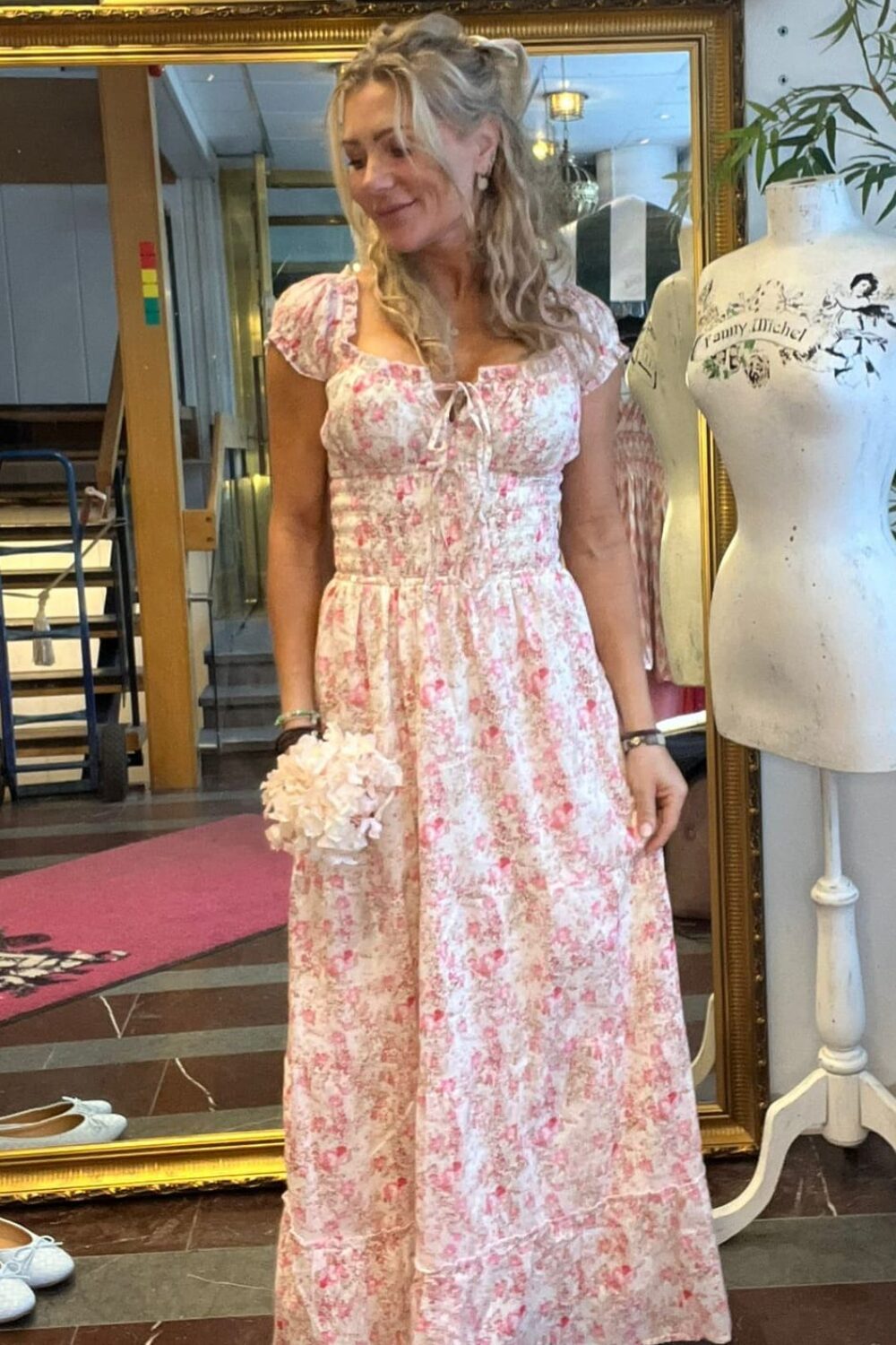 Kvinna i ljusrosa, blommig långklänning står och ler lätt medan hon håller en liten bukett. Hon står framför en stor guldramsspegel i en ateljé/butik med inramade foton och provdocka med texten ”Fanny Michel”.