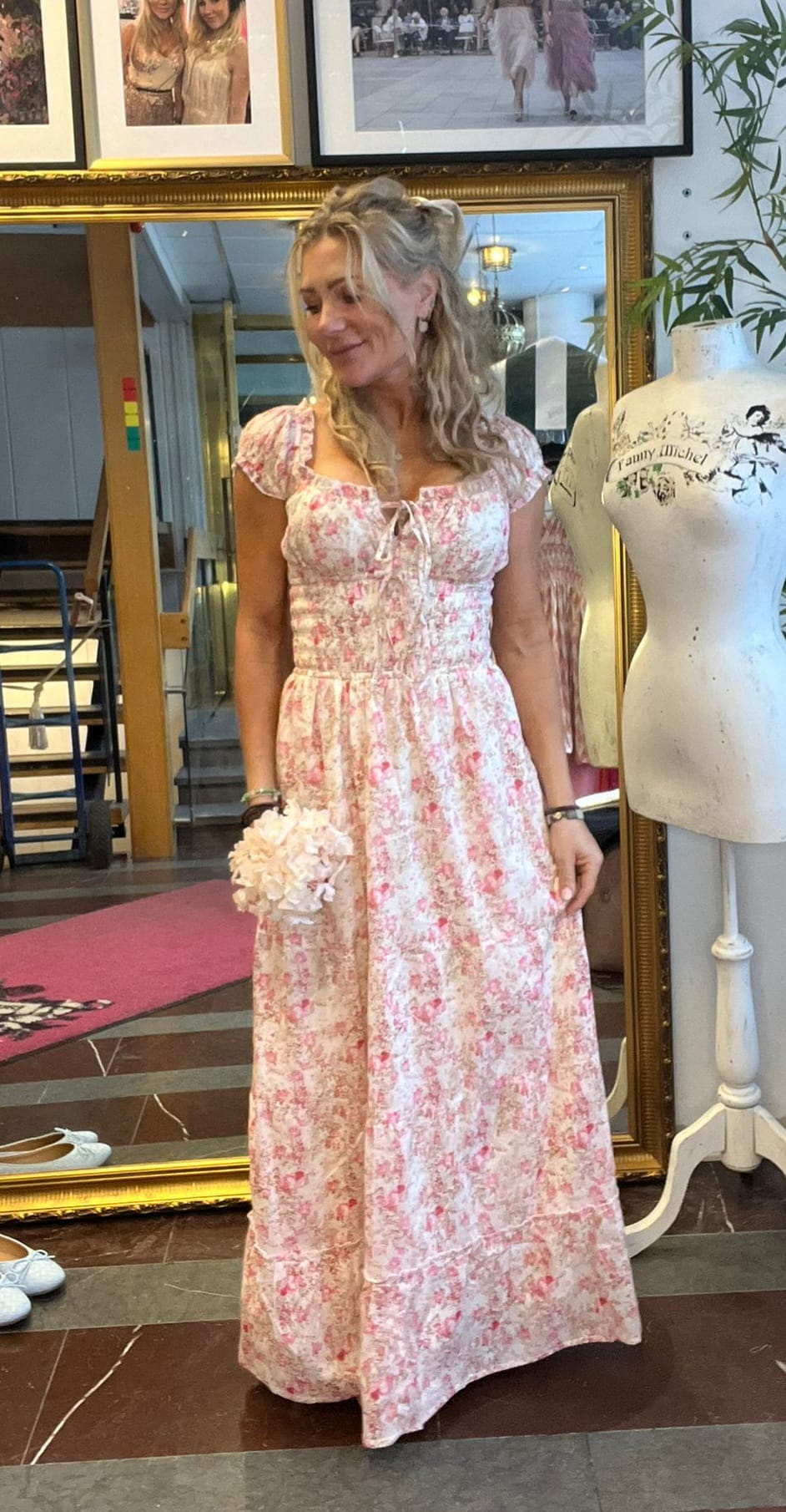 Kvinna i ljusrosa, blommig långklänning står och ler lätt medan hon håller en liten bukett. Hon står framför en stor guldramsspegel i en ateljé/butik med inramade foton och provdocka med texten ”Fanny Michel”.