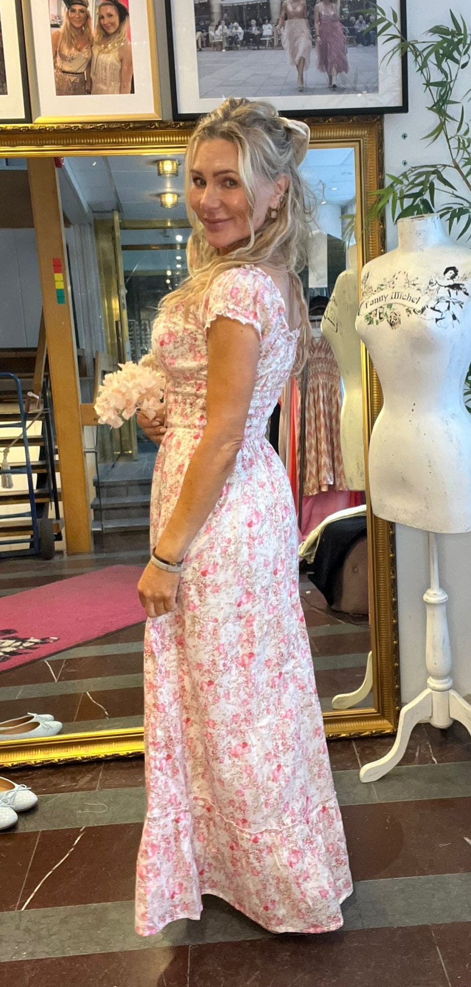 Kvinna i lång rosa‑vit blommig klänning står i profil och ler mot kameran, med en ljus bukett i handen. Hon är i en klädbutik/ateljé bredvid provdocka med texten “Tammy Milchel” och inramade foton.