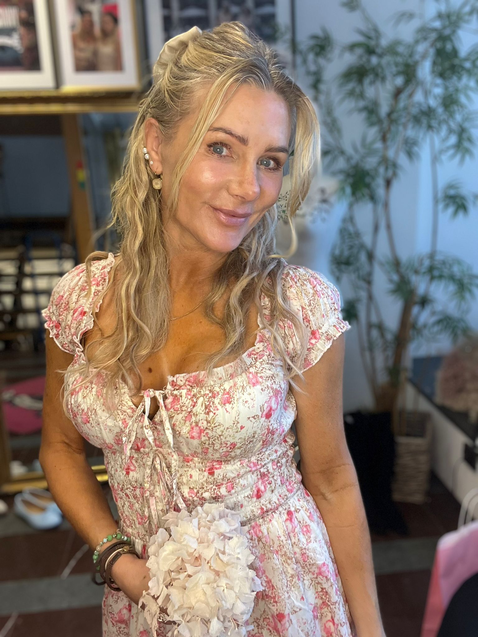 Leende blond kvinna poserar och håller en vit blomsterbukett, klädd i en rosa- och vitblommig klänning med knyt fram. Inomhusmiljö med spegel, foton i bakgrunden och en stor grön växt.