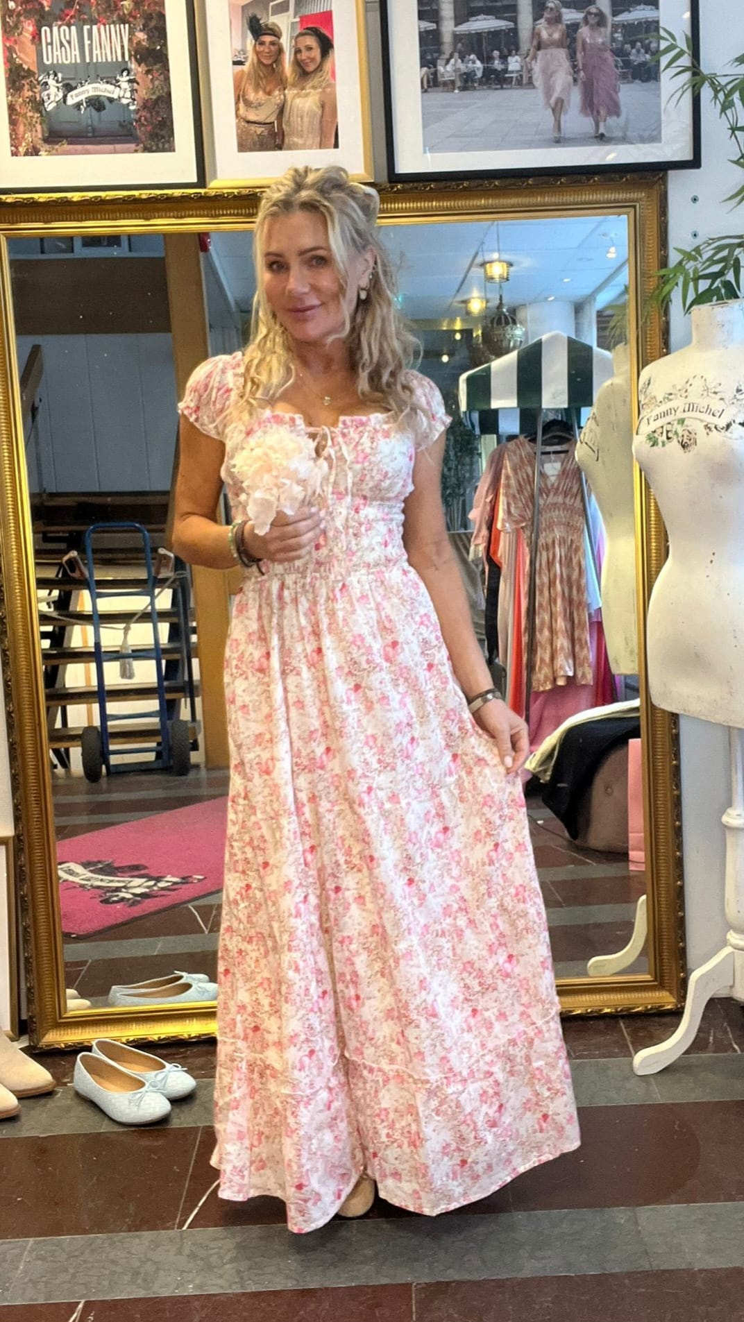 Kvinna står framför en stor guldramsspegel och håller en blomma, iklädd lång rosa blommig klänning med puffärmar. I bakgrunden syns klädbutik med klänningar, provdocka och inramade foton; text: ”CASA FANNY”.